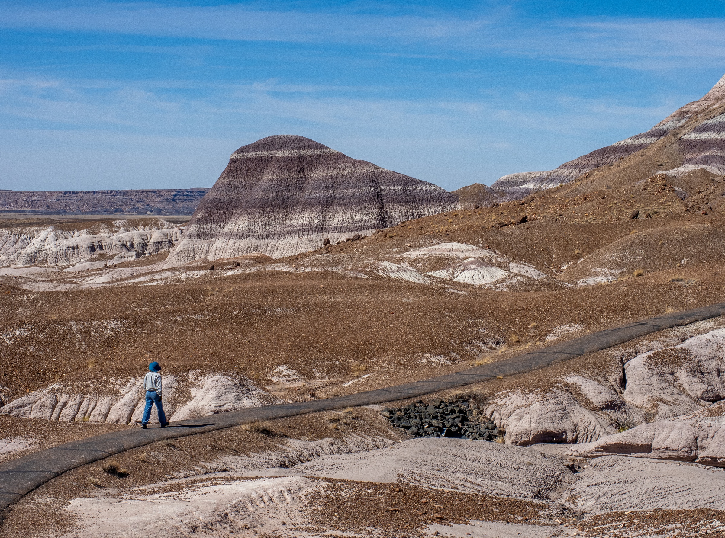 A person walks along a road through layered, colorful badlands under a clear blue sky.