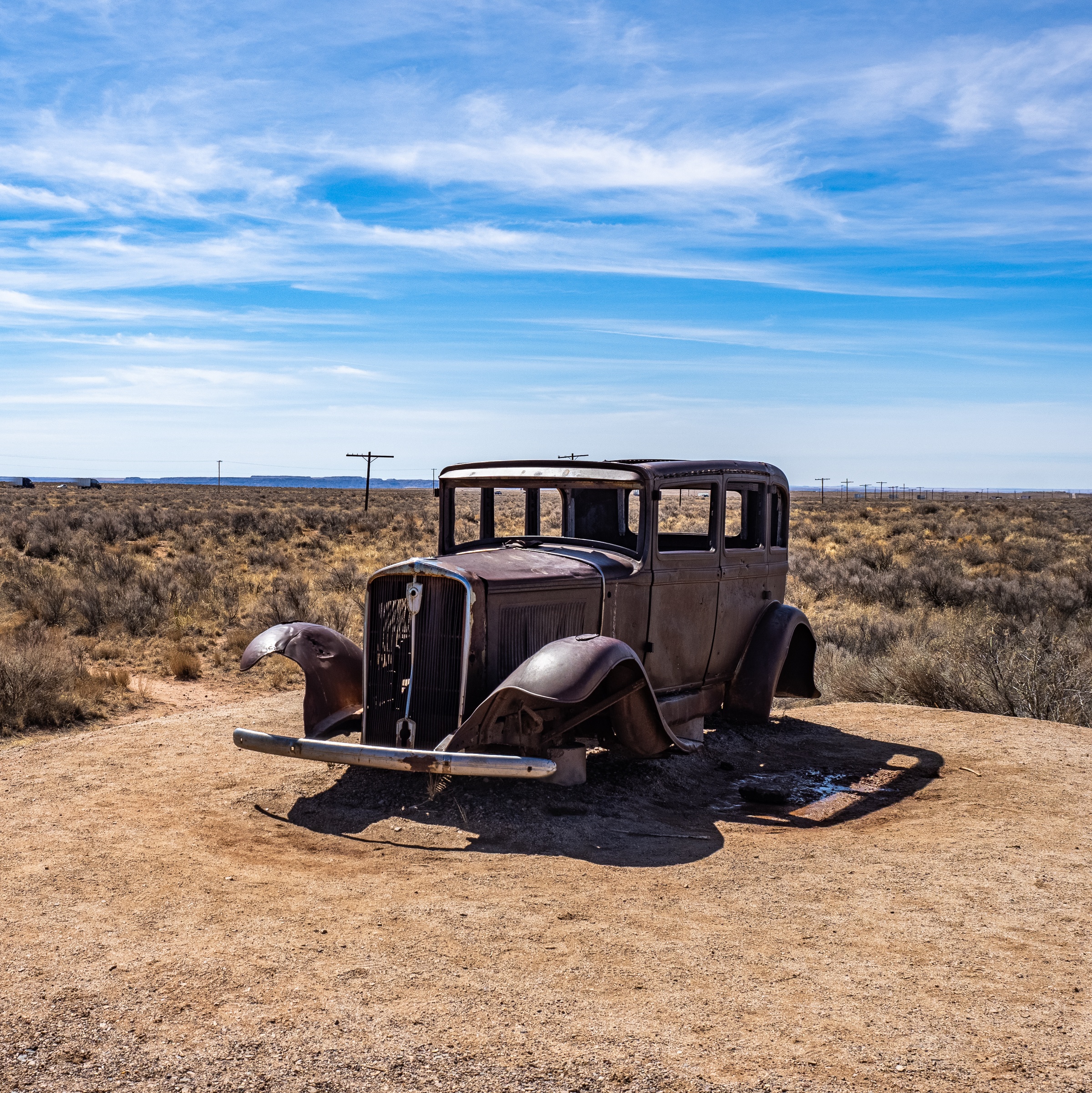 A rusted vintage car sits on a small rise within a vast, arid landscape under a blue sky.