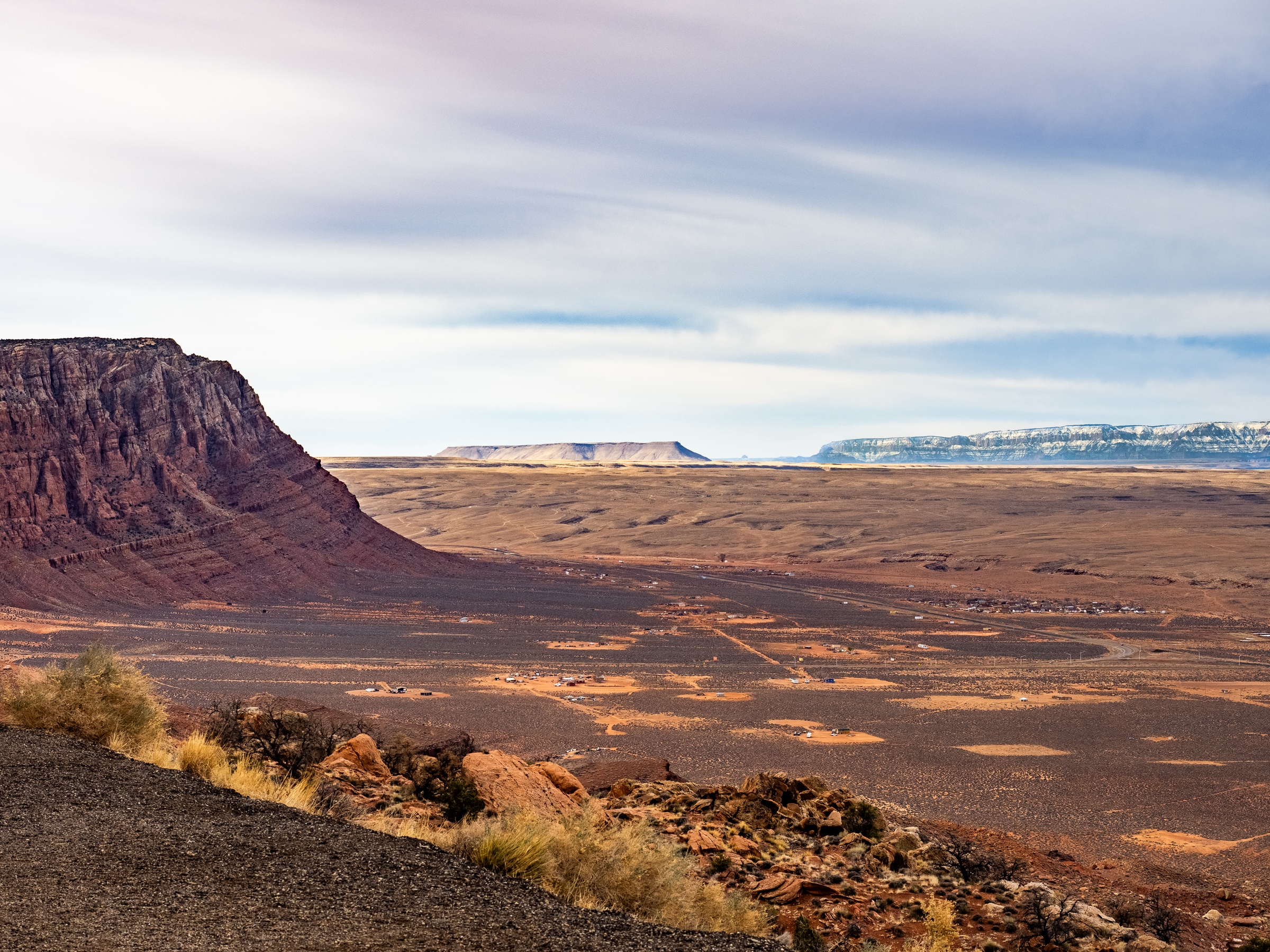 Red rock mesa overlooks a vast, flat desert landscape under cloudy skies.