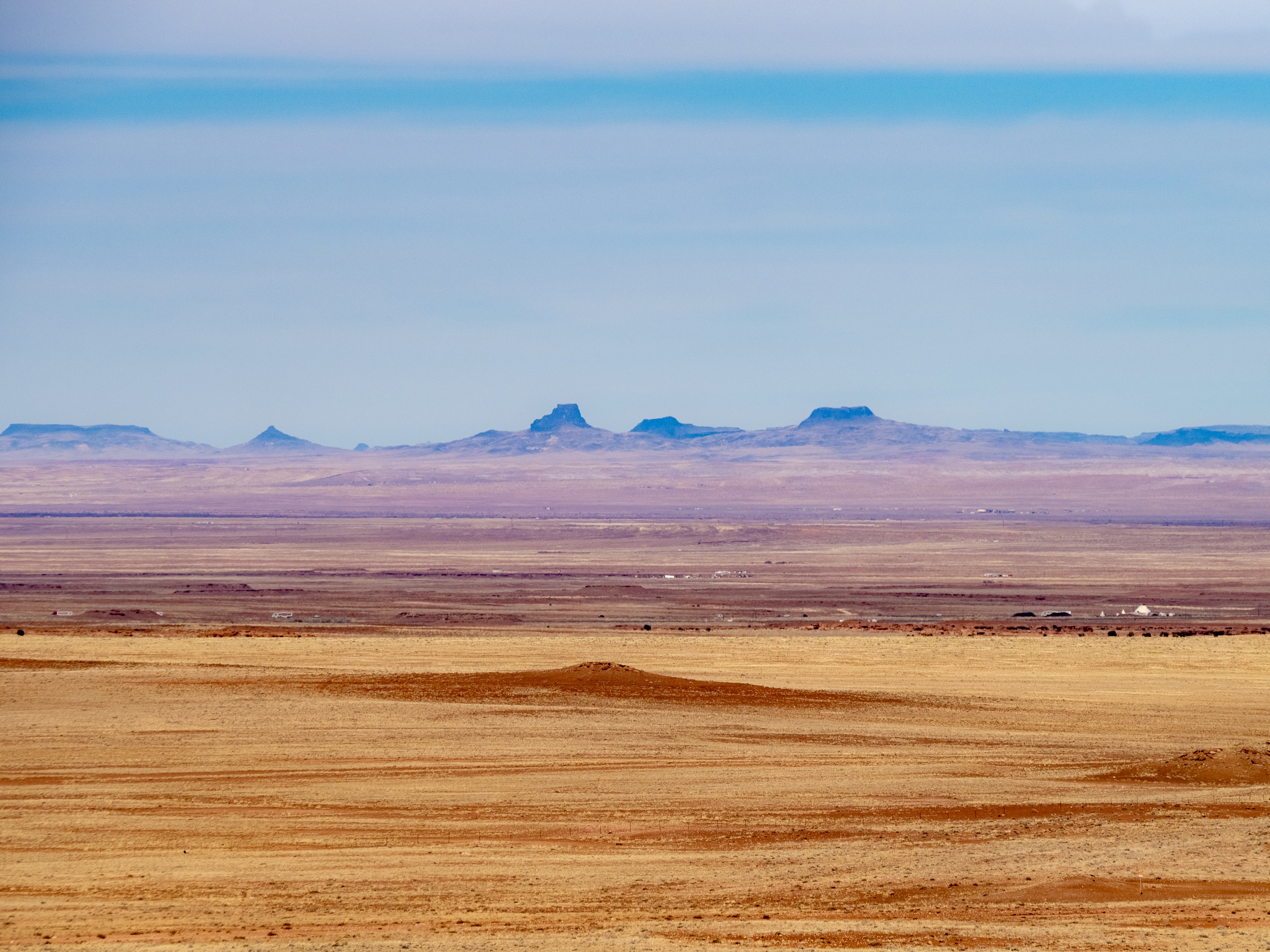 Vast tan desert stretches to a distant range of mesas under a pale blue sky.