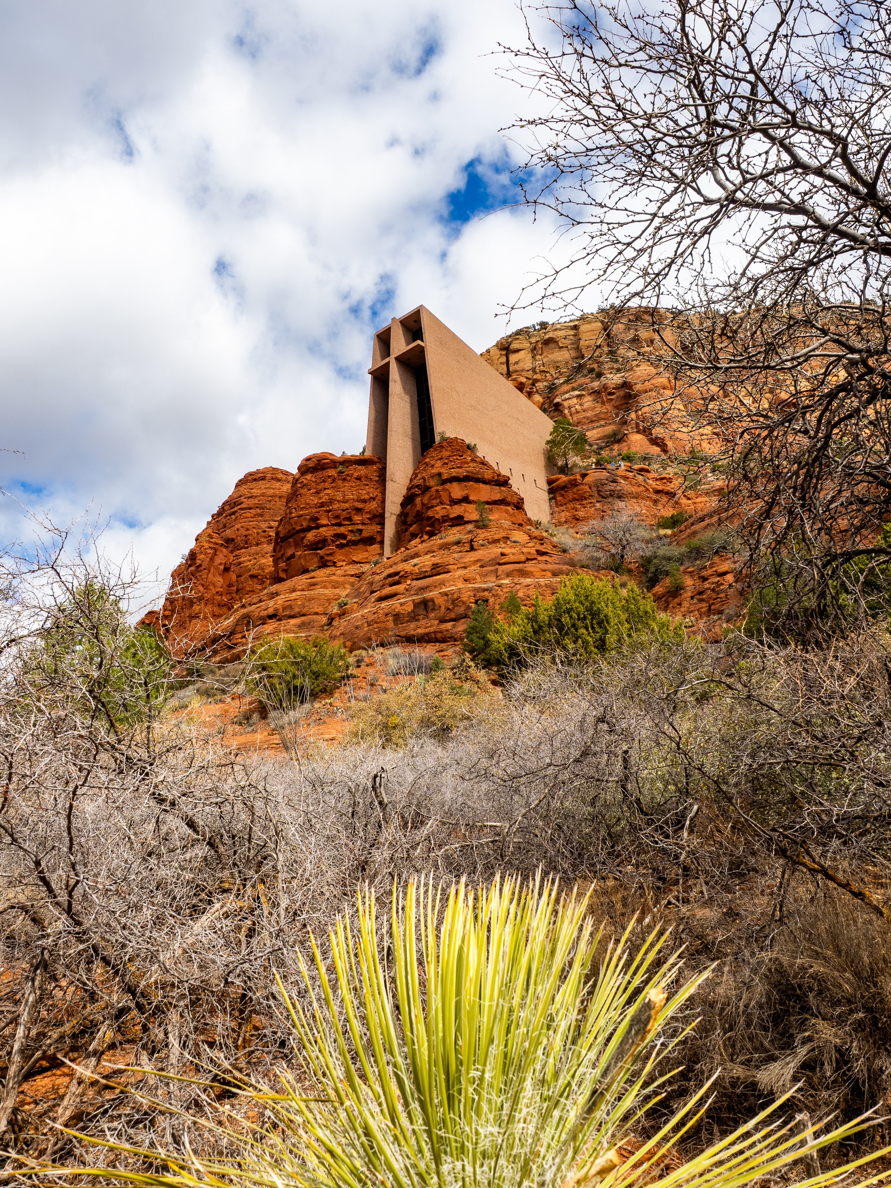 Chapel of the Holy Cross sits atop a red rock butte, framed by desert vegetation and cloudy skies.
