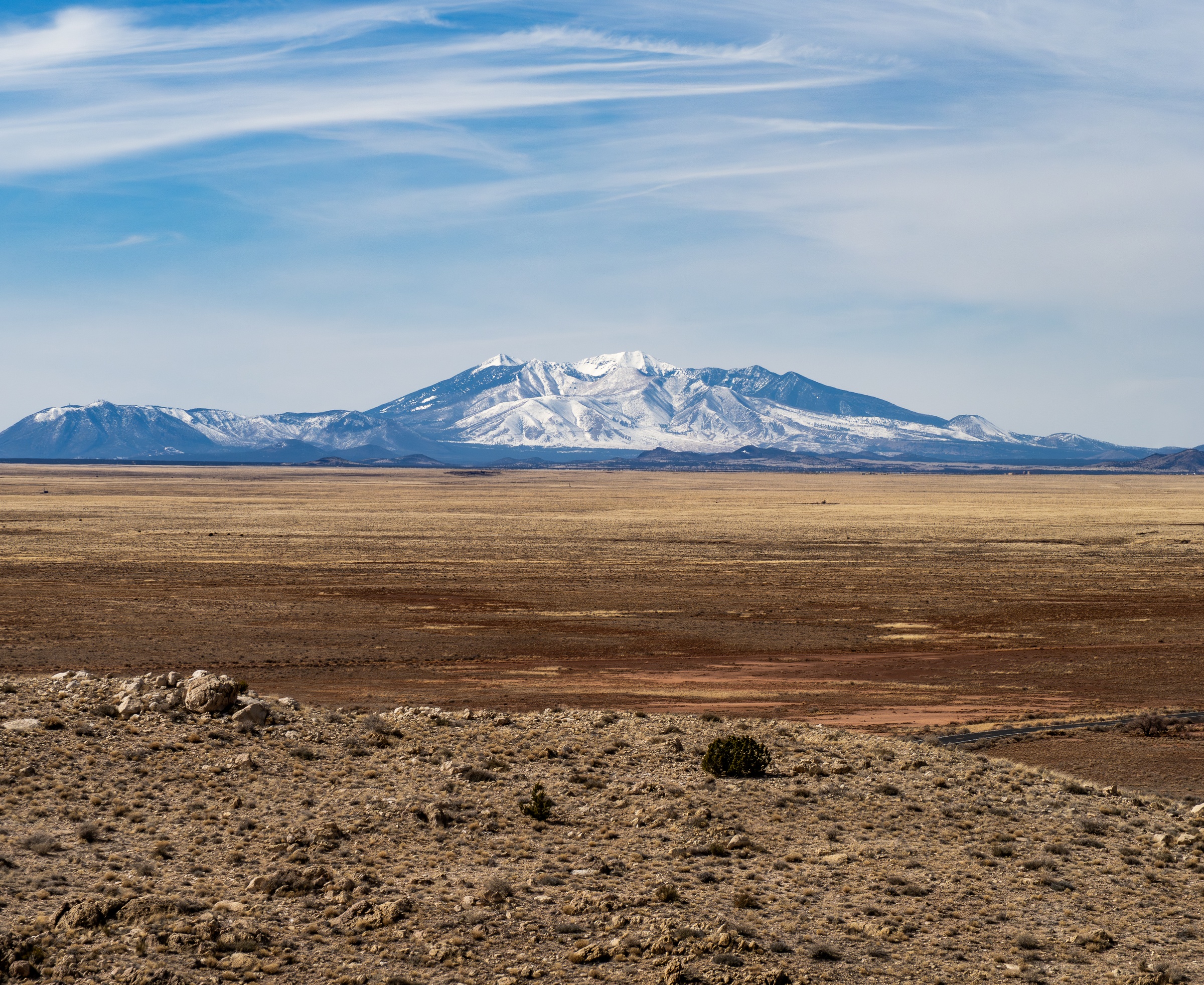 A snow-capped mountain range rises above a vast, dry, brown landscape under a blue sky.