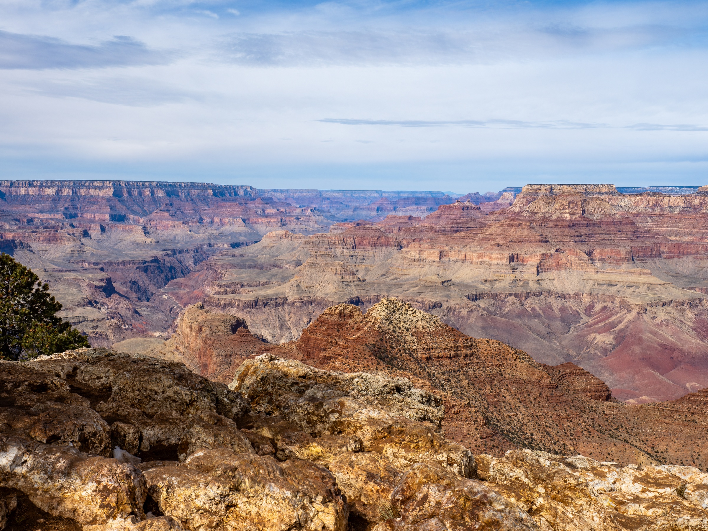 Vast layered rock formations and canyons stretch across a landscape under a cloudy sky.