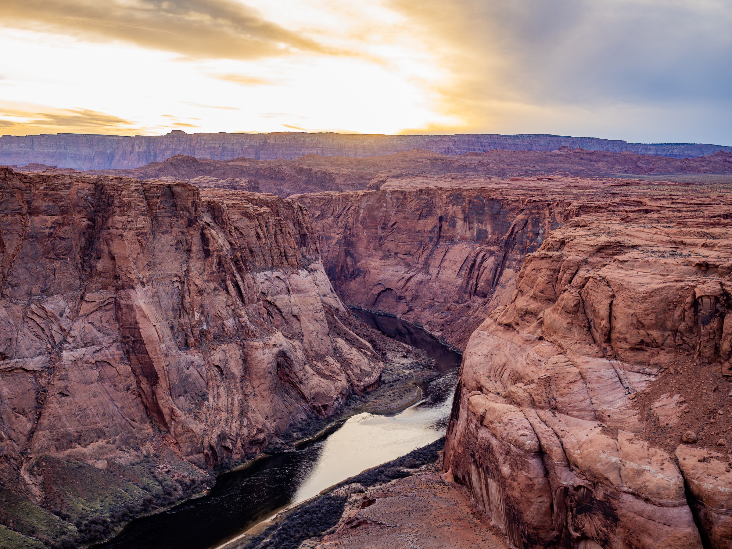 A river winds through a vast canyon of layered red rock under a warm sunset sky.