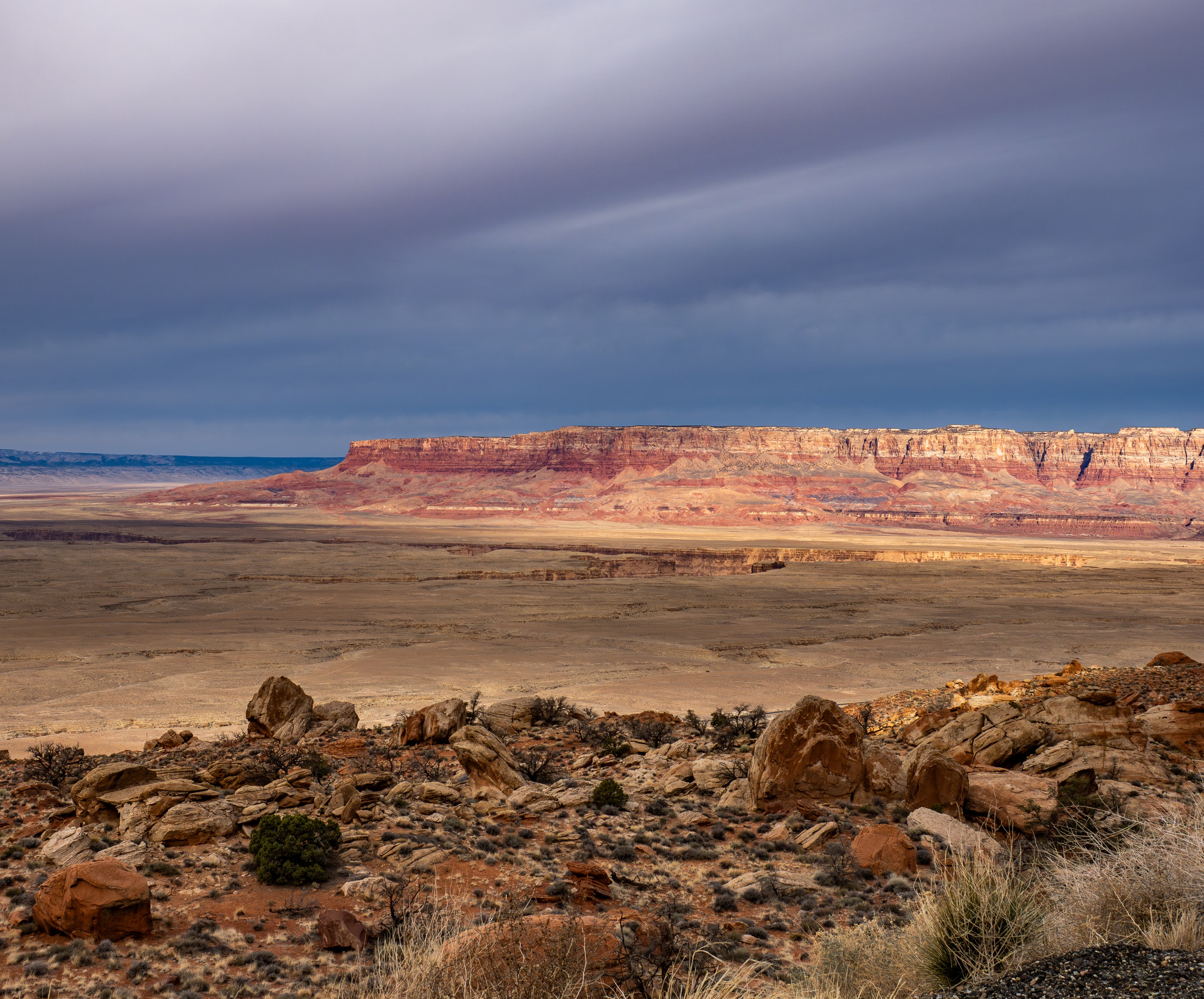 Red rock cliffs rise above a vast, dry landscape under an overcast sky.