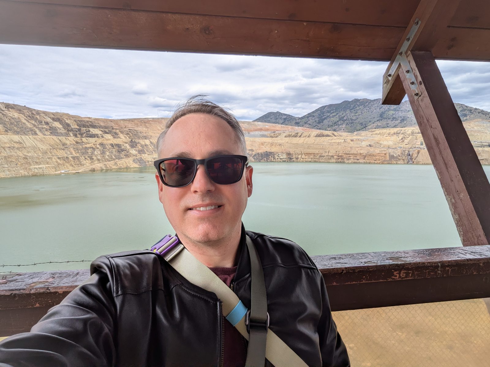 A man in a black leather jacket and sunglasses takes a selfie in front of a blue pond.