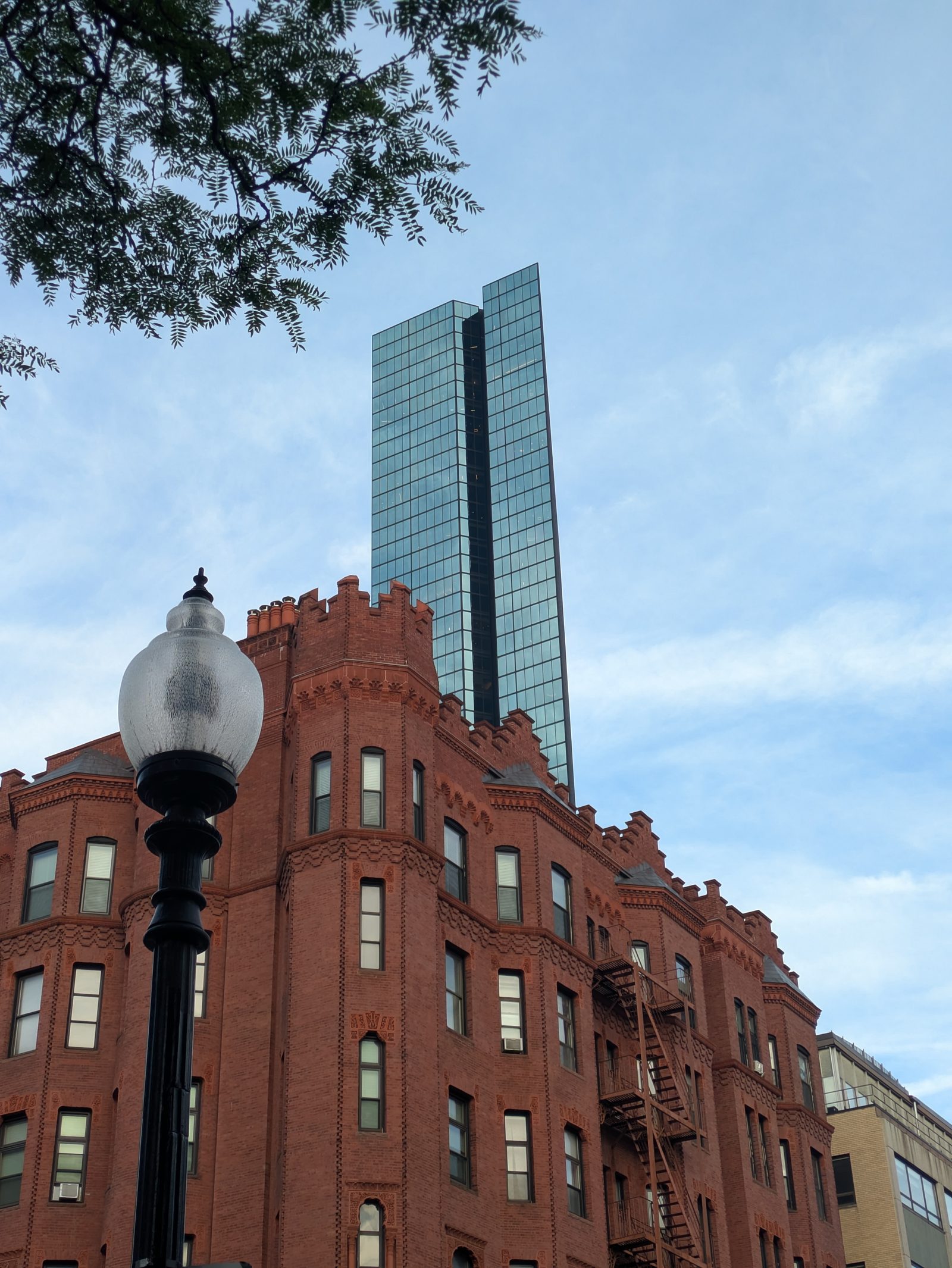 A glass skyscraper rises behind an old brick building against a light blue summer sky