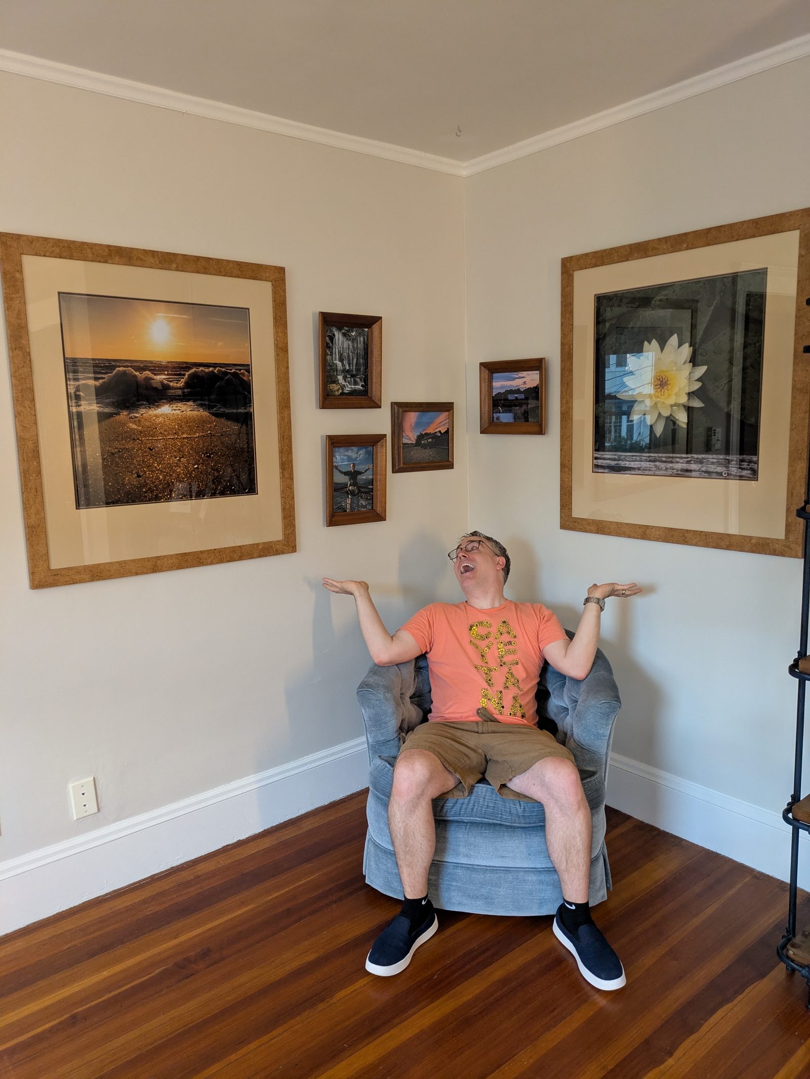 A man sits in a blue chair with a funny expression seated in front of two walls in a corner with large photos on the wall.