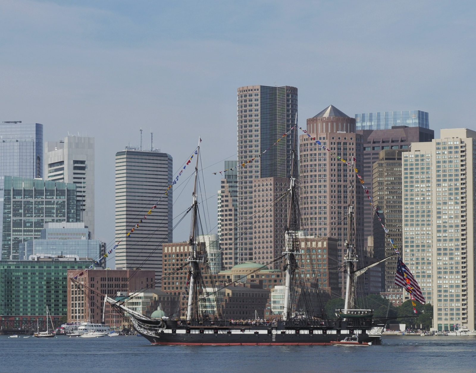A large sailing ship with multiple masts floats in a harbor before a city skyline.
