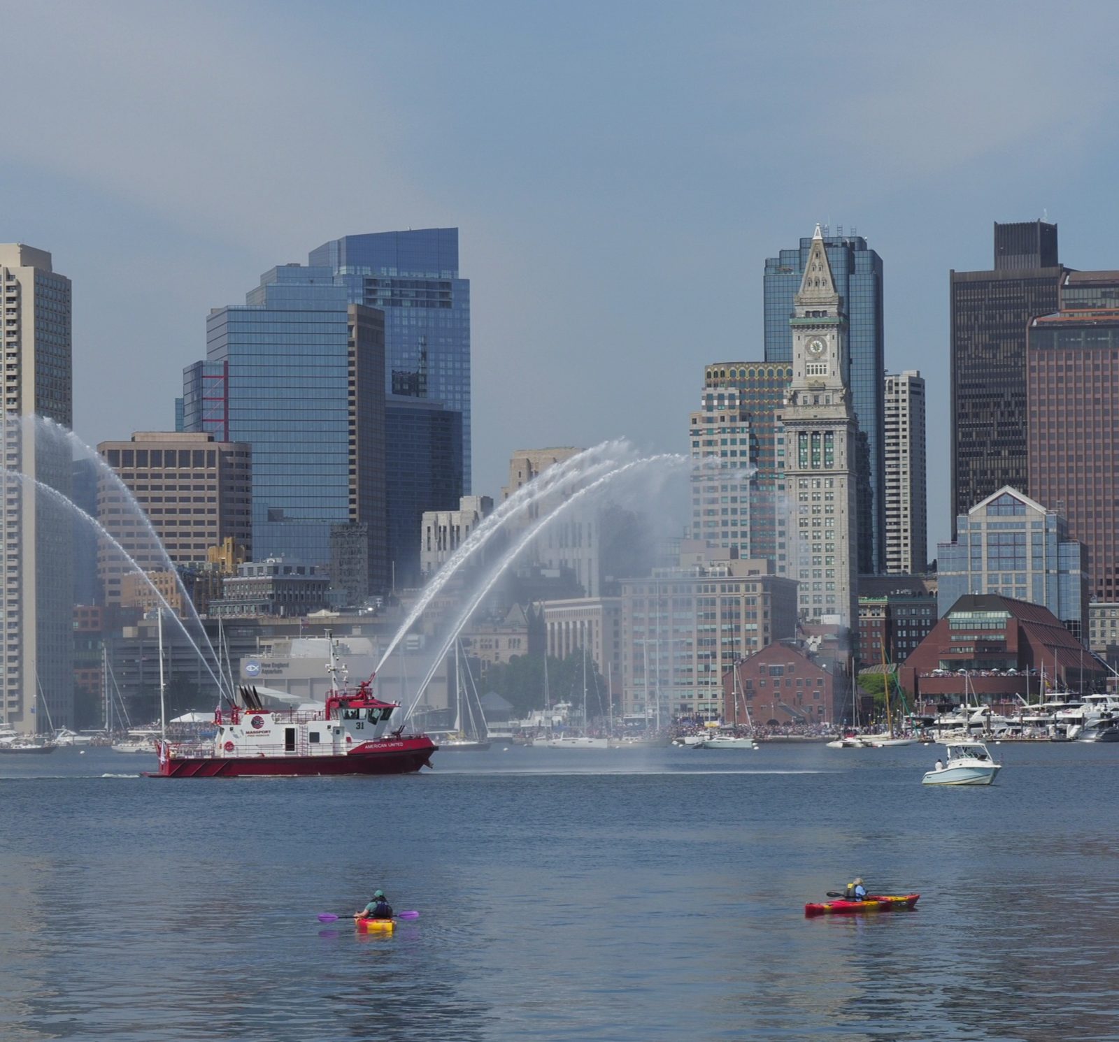 A fireboat sprays water arcs in a harbor against a city skyline with skyscrapers.