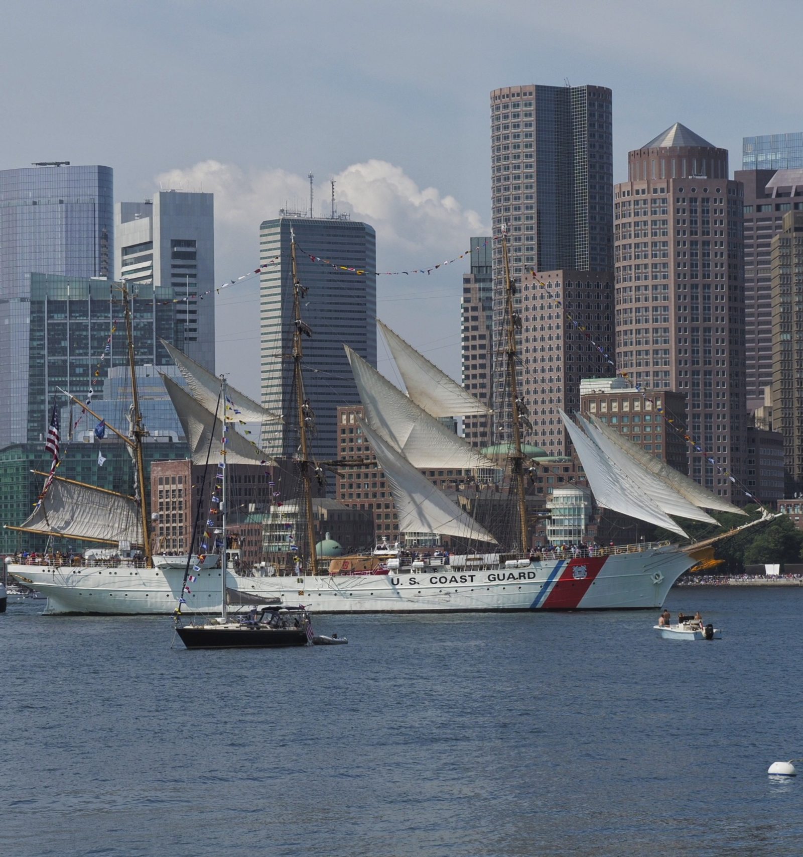 A U.S. Coast Guard sailing ship and smaller boats float before a city skyline of skyscrapers