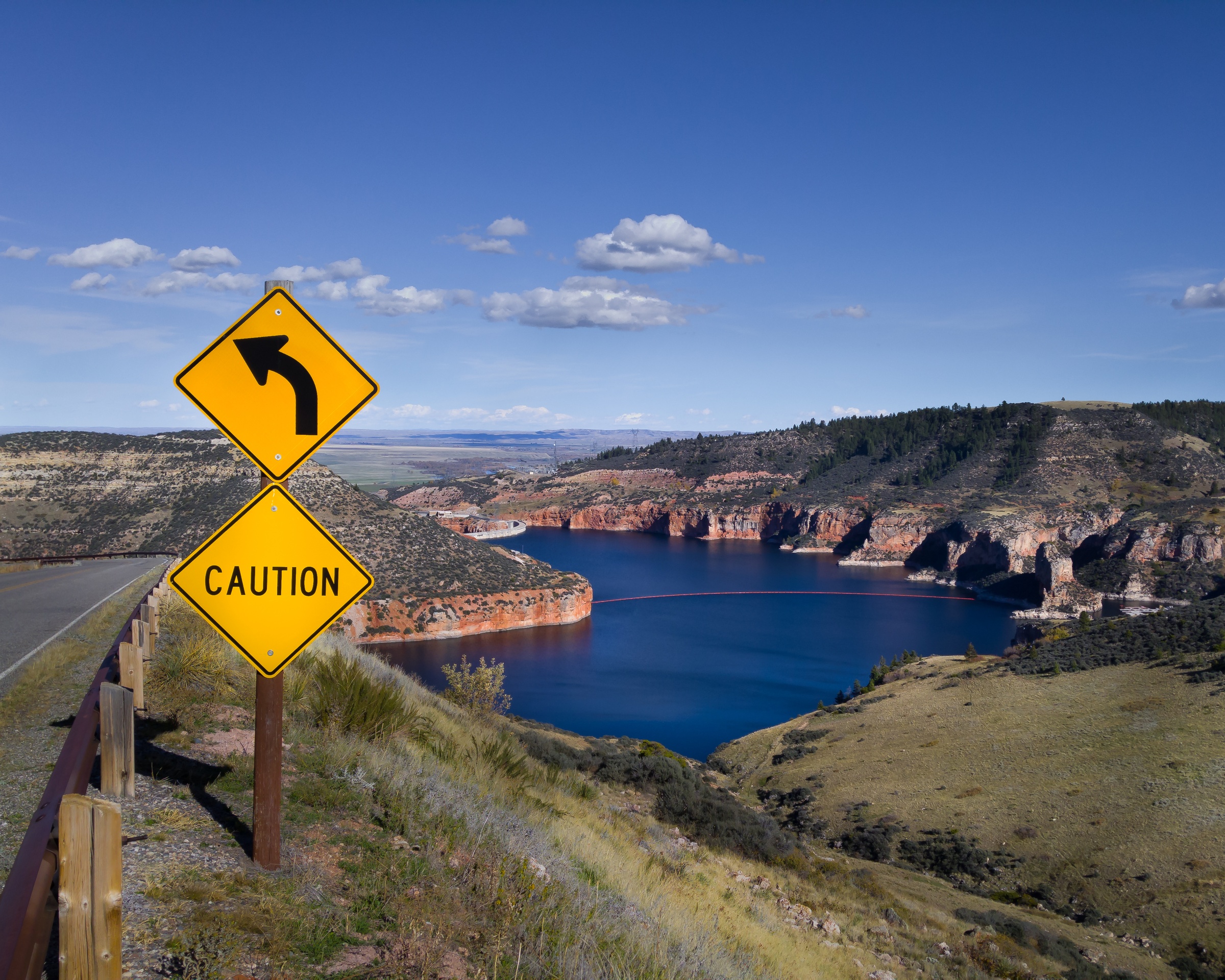 A yellow caution sign warns of a sharp curve overlooking a scenic lake and layered rock formations.