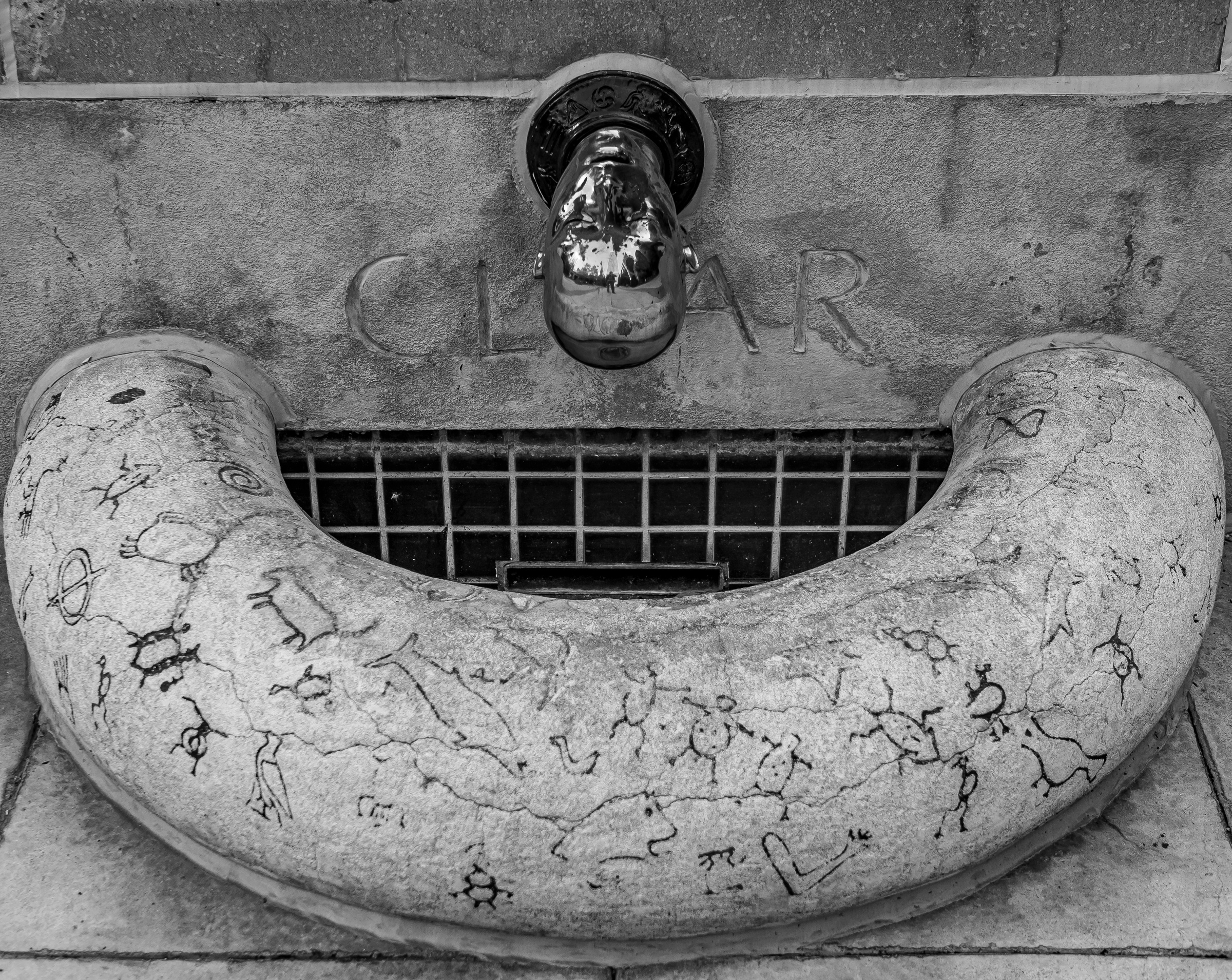 A weathered stone drinking fountain with carved markings sits on a textured concrete surface.