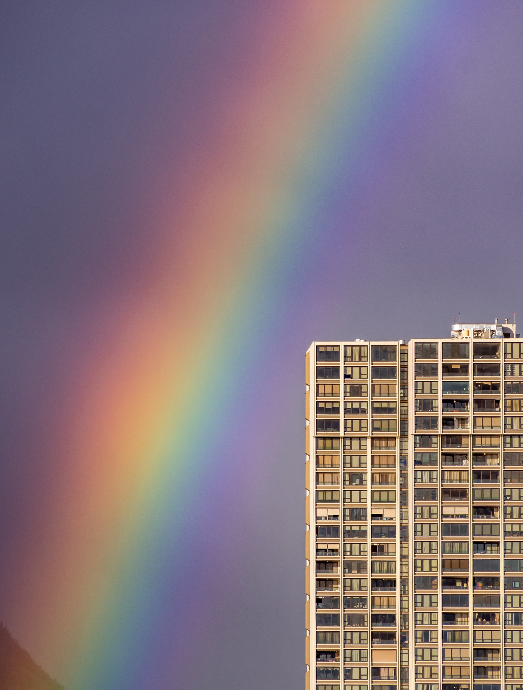 A vibrant rainbow arcs over a tall apartment building against a cloudy sky.