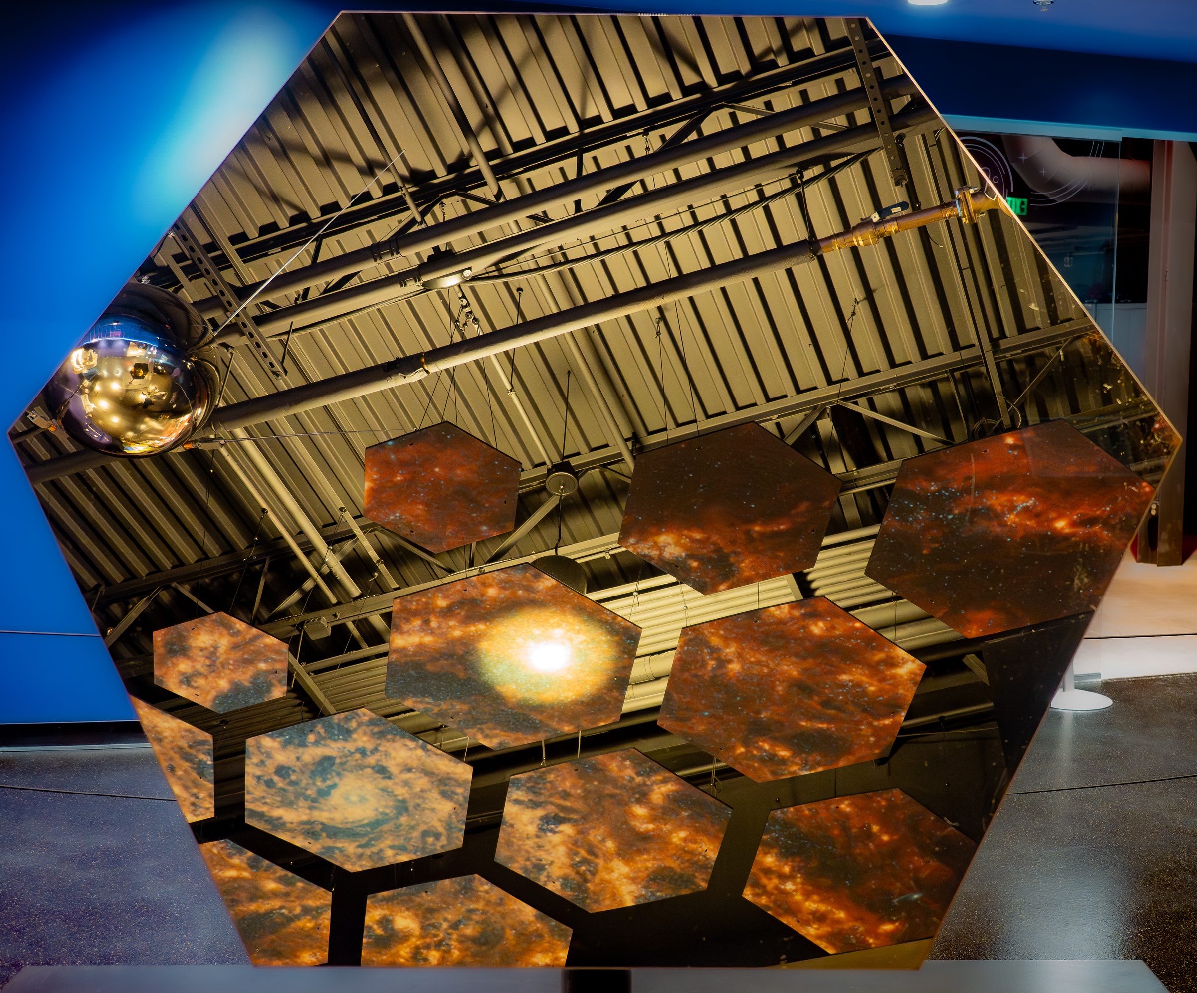 A large hexagonal mirror displays images of space and a reflective silver sphere against a ceiling structure.