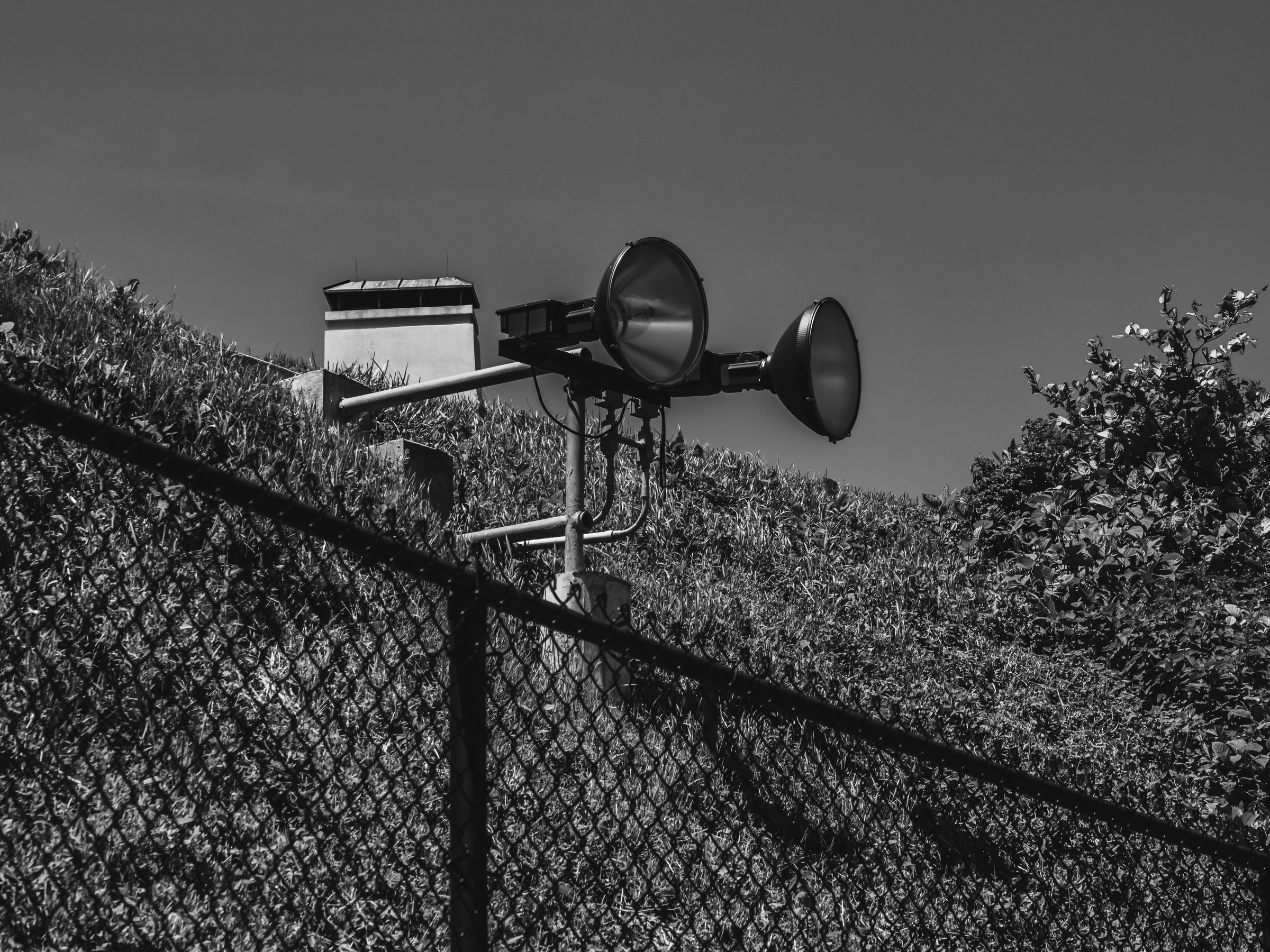 Spotlights extend from a metal structure above a chain-link fence and overgrown vegetation.