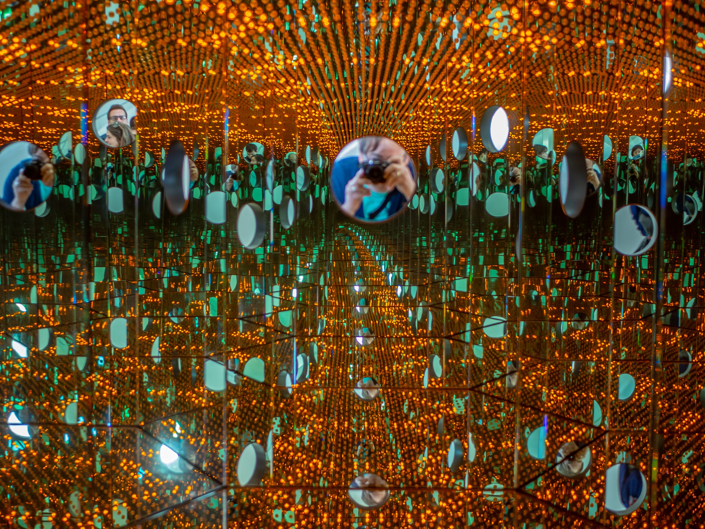 A person takes a photo within an immersive mirrored room filled with orange lights and circular reflections.