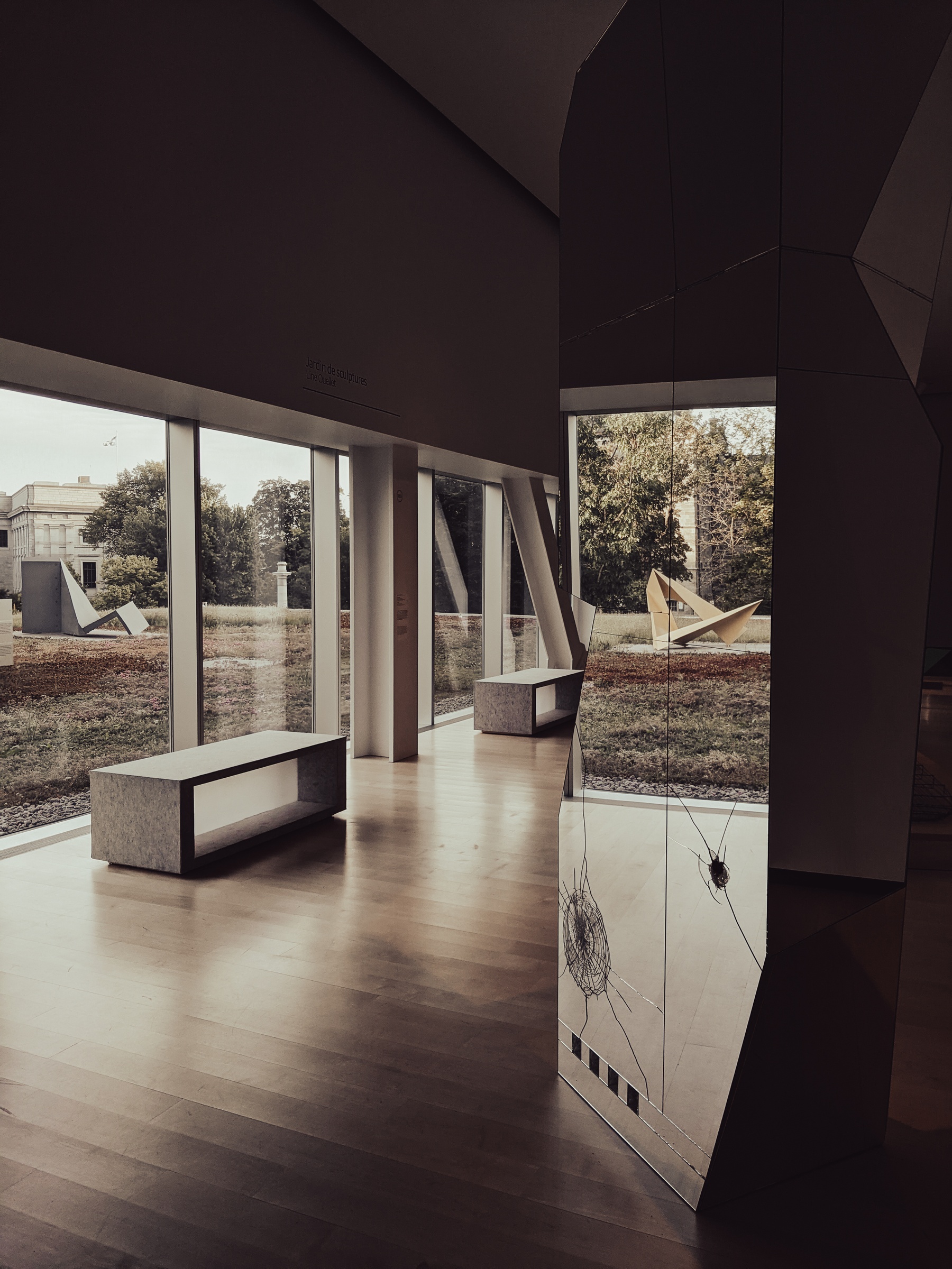 A modern art museum interior features wood floors, benches, and large windows overlooking outdoor sculptures.
