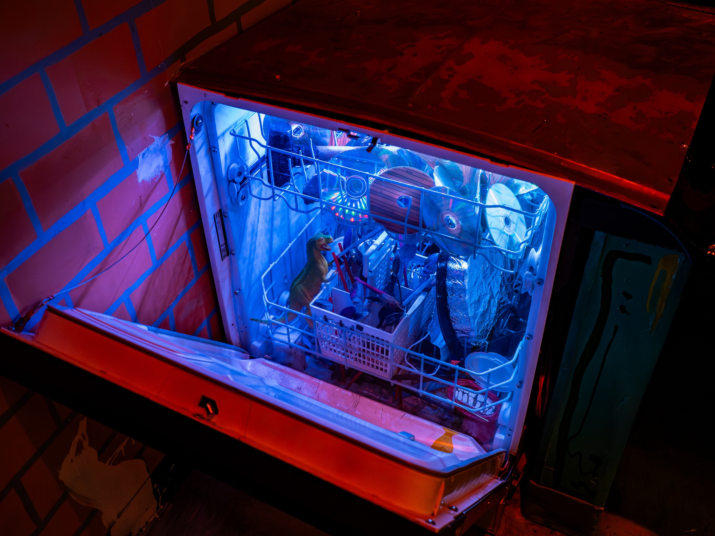 An open dishwasher glows with blue light, containing a small dog and various objects against a brick wall.