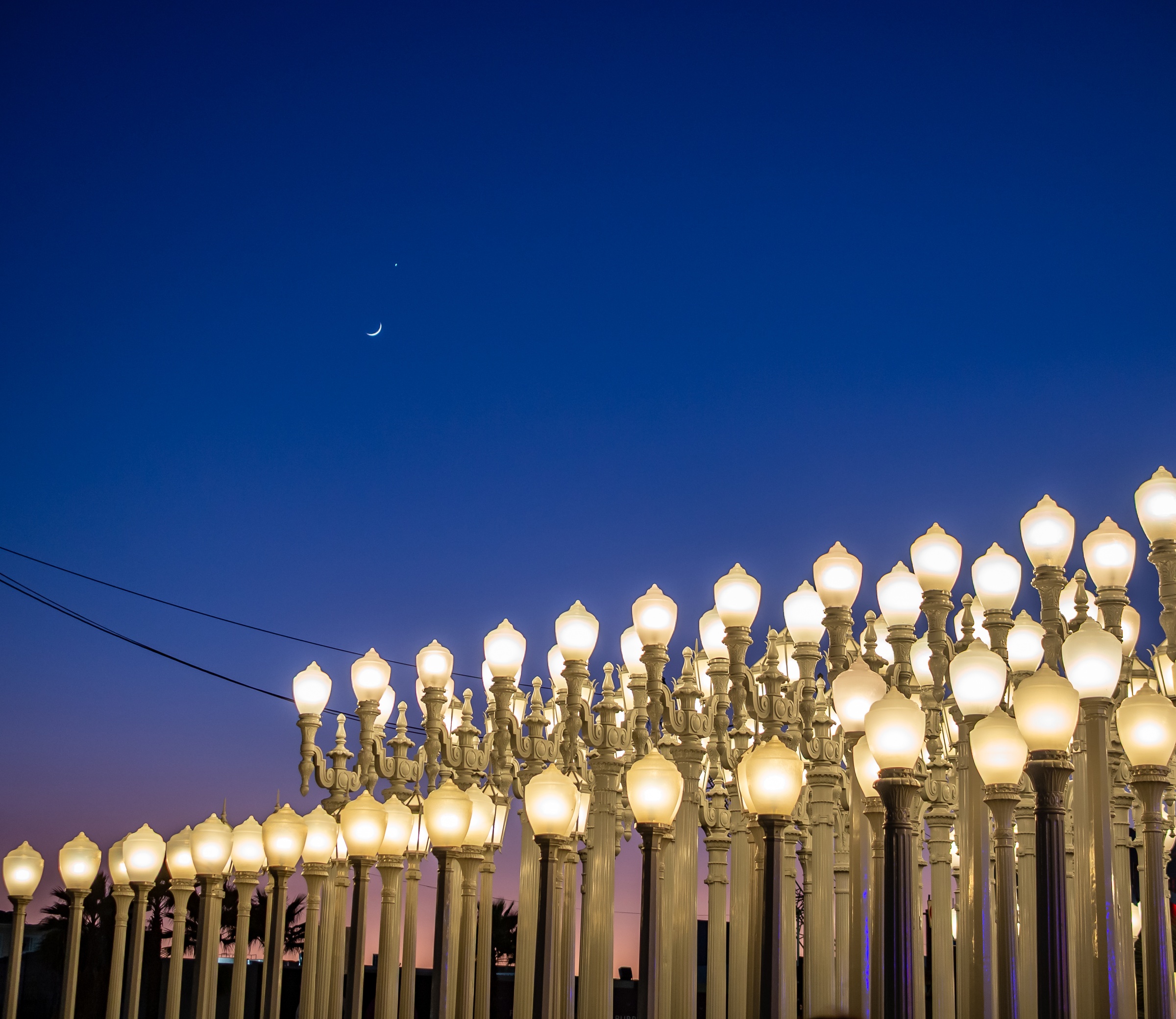 Rows of illuminated street lamps stretch toward a twilight sky with a crescent moon.