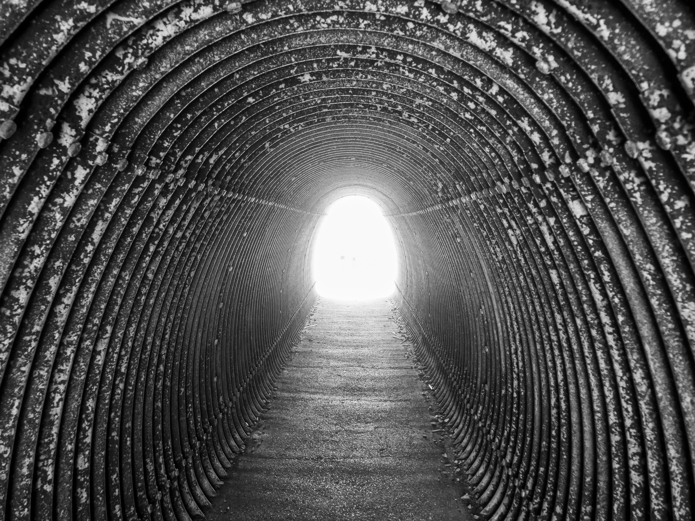 A black and white photograph shows a tunnel with corrugated metal walls leading to bright light.