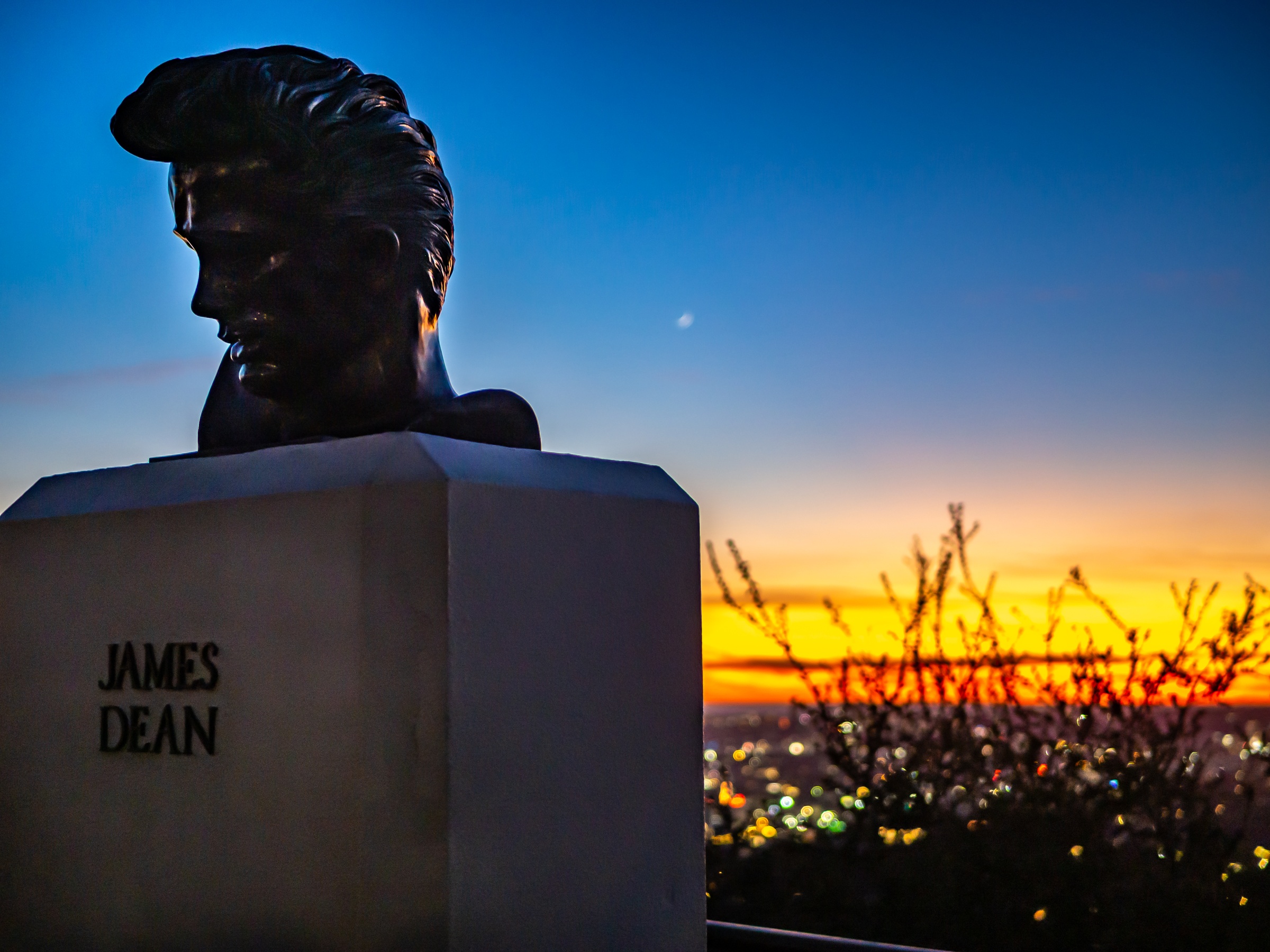 A bronze bust of James Dean sits on a pedestal against an orange and blue sunset cityscape.