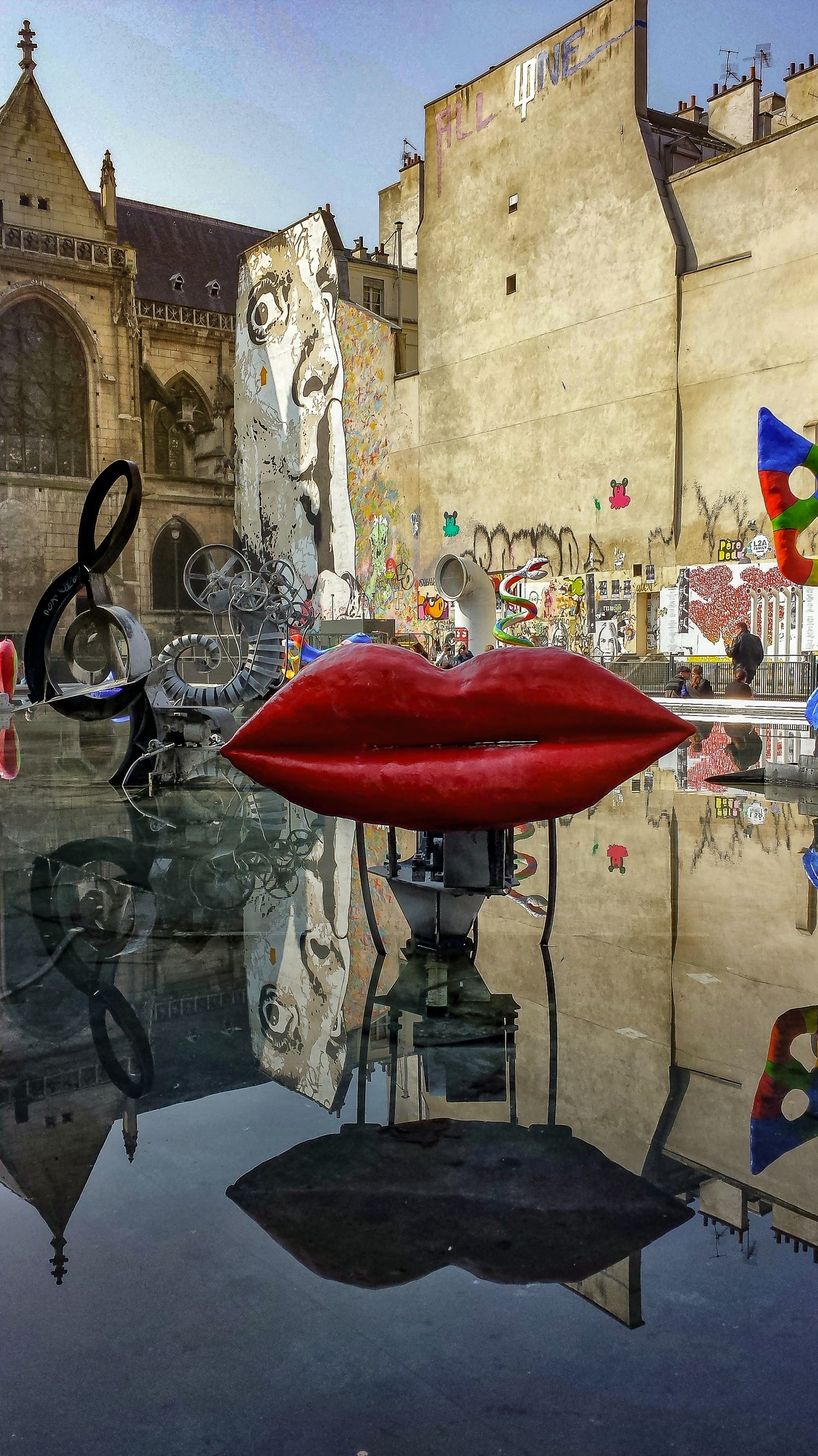 Red lips and other sculptures reflect in a pool of water before a building with street art.