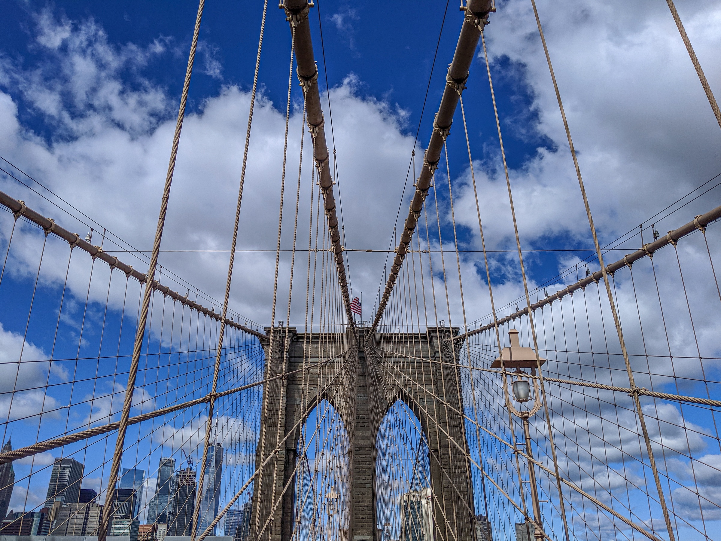 The Brooklyn Bridge arches against a bright blue sky with fluffy white clouds above the New York City skyline.