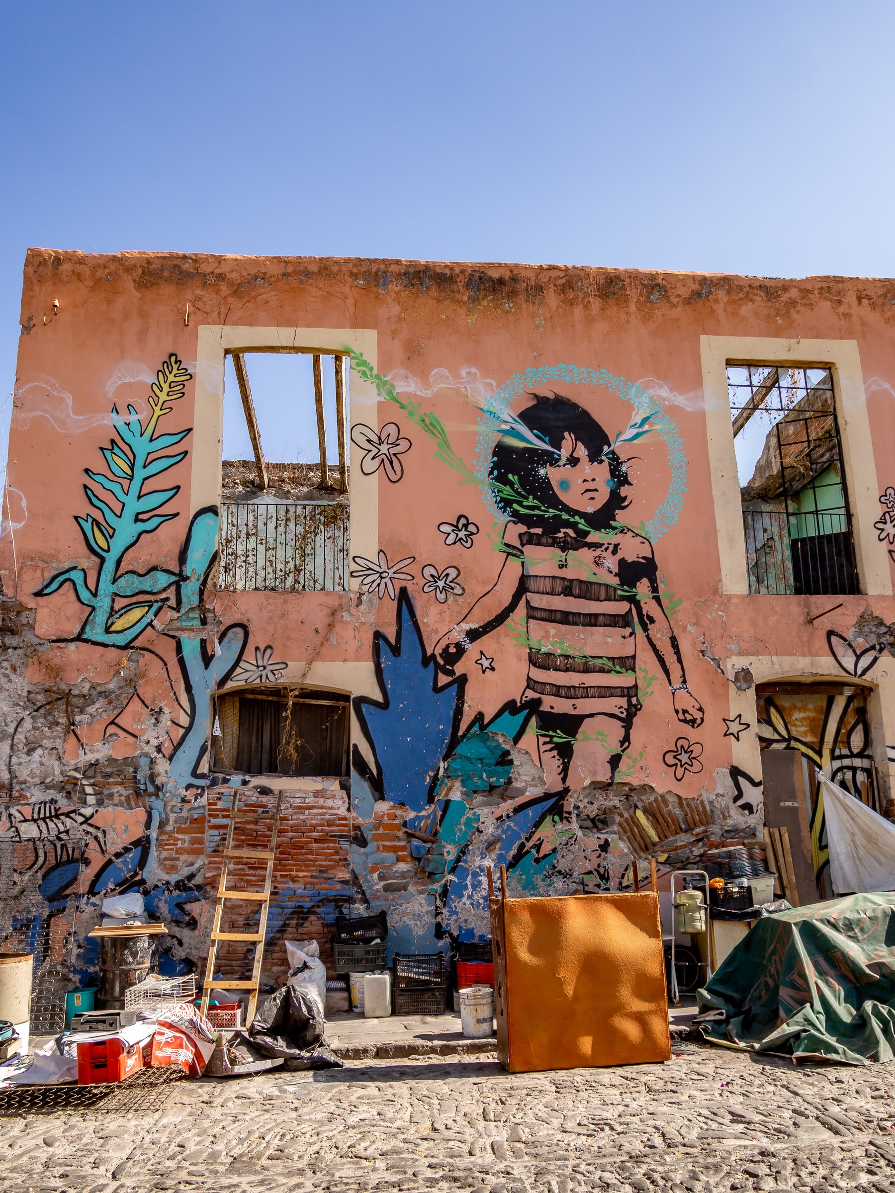 A black and white mural of a child adorns a weathered, orange building with surrounding graffiti and debris.