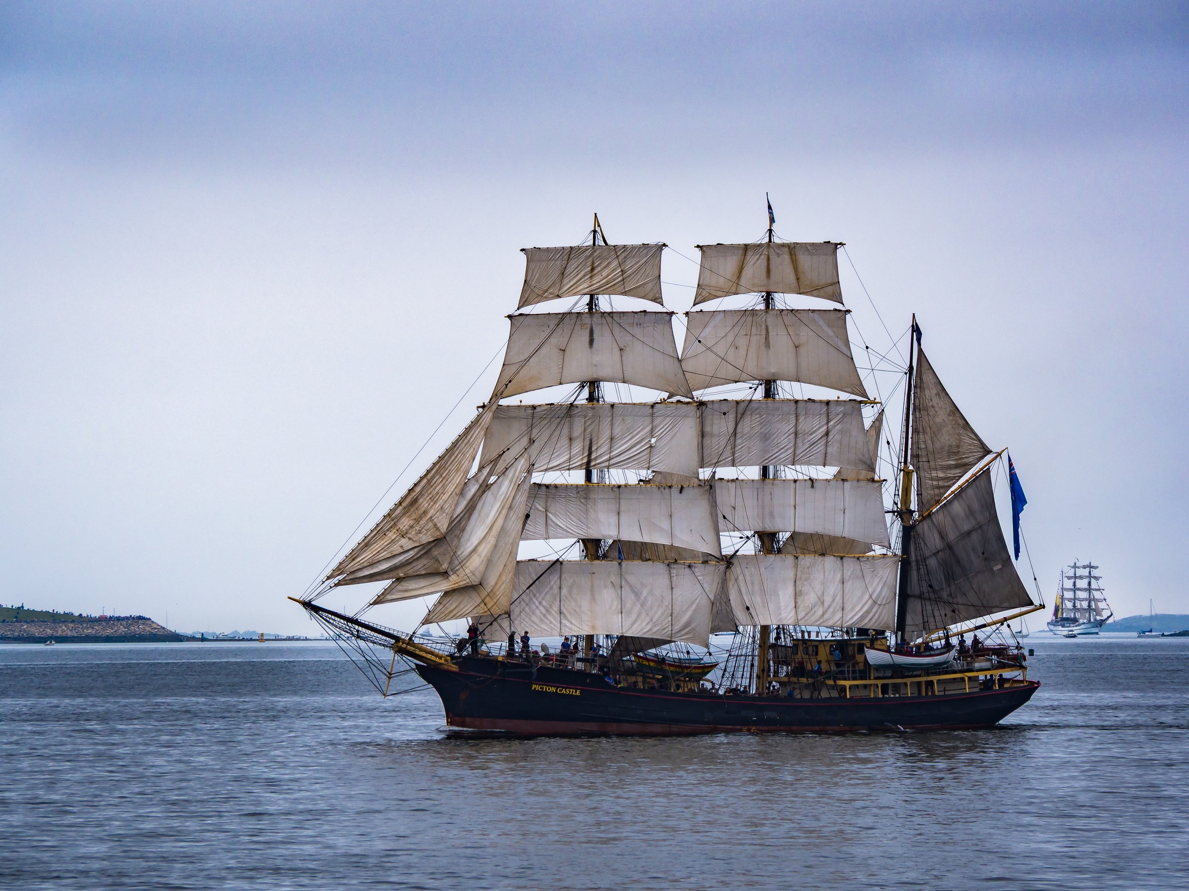 A tall sailing ship with multiple sails floats on a gray body of water under an overcast sky.