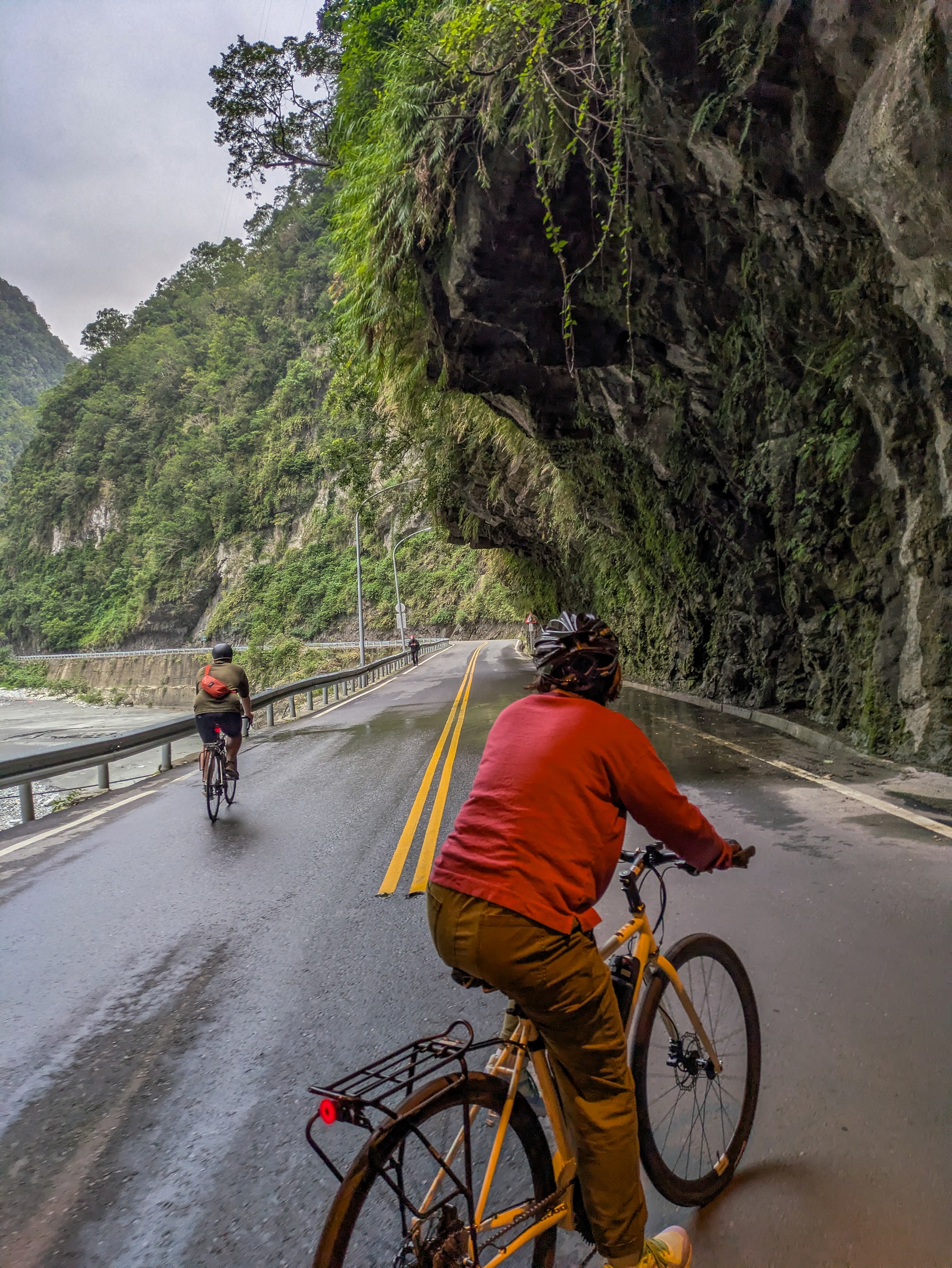 Two cyclists ride along a wet road beside a steep, mossy cliff face.