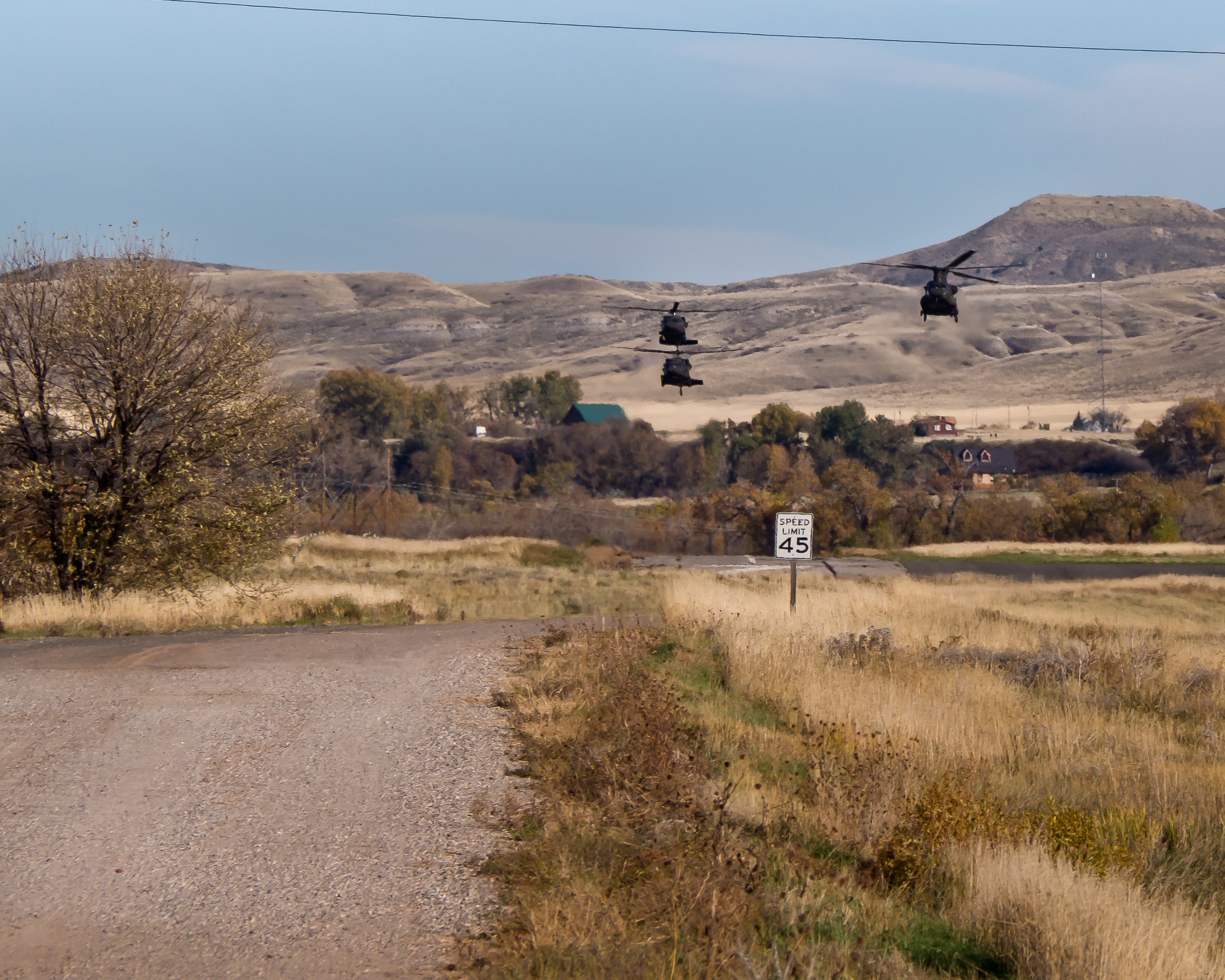 Three helicopters fly over a rural landscape with fields, trees, and a road with a speed limit sign.