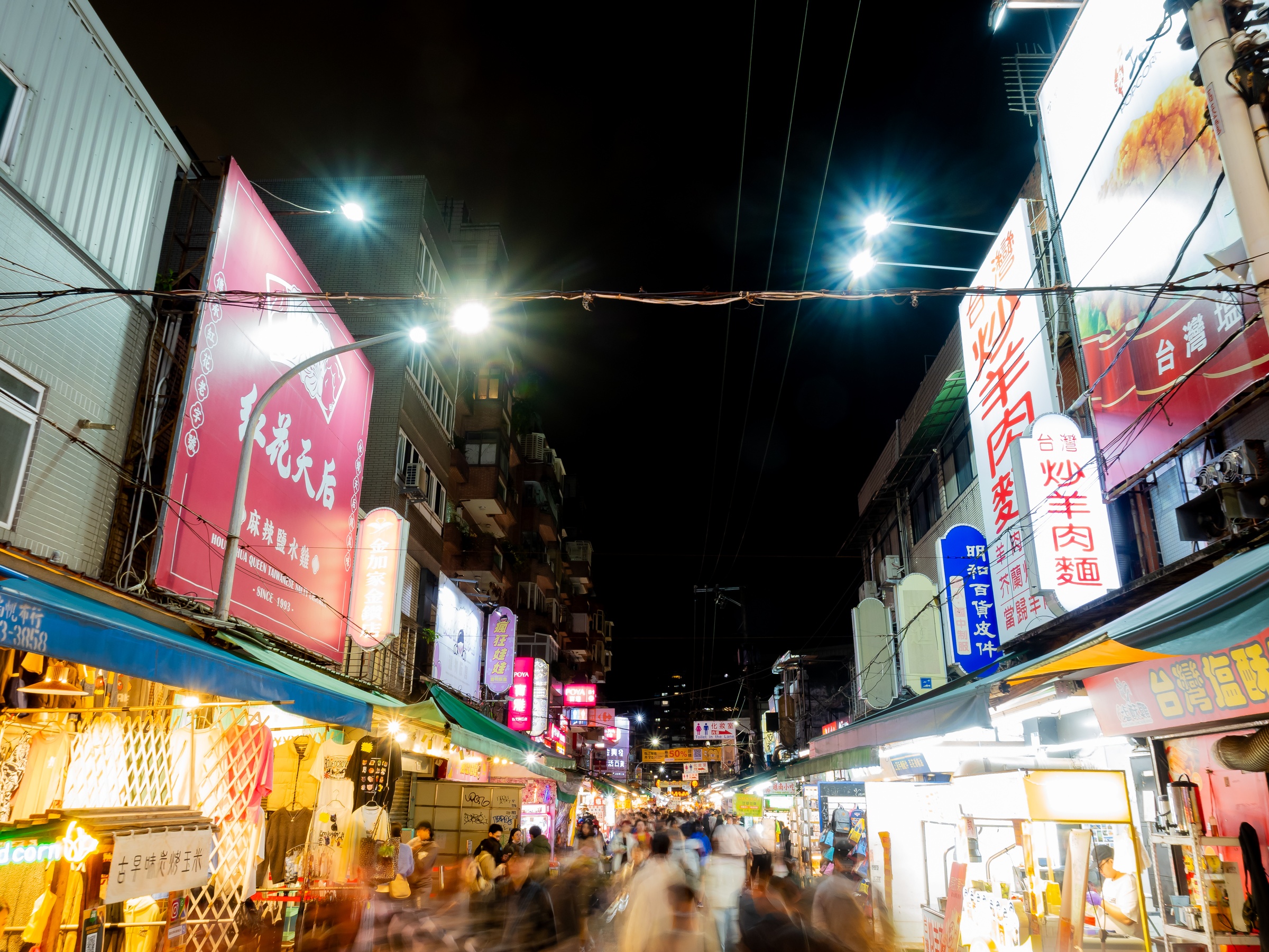 A bustling night market street in Taiwan is filled with brightly lit signs and blurred pedestrians.