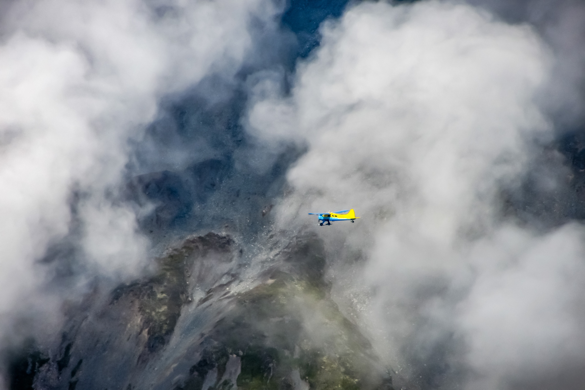 A yellow airplane flies through a cloudy sky above a mountainous landscape.