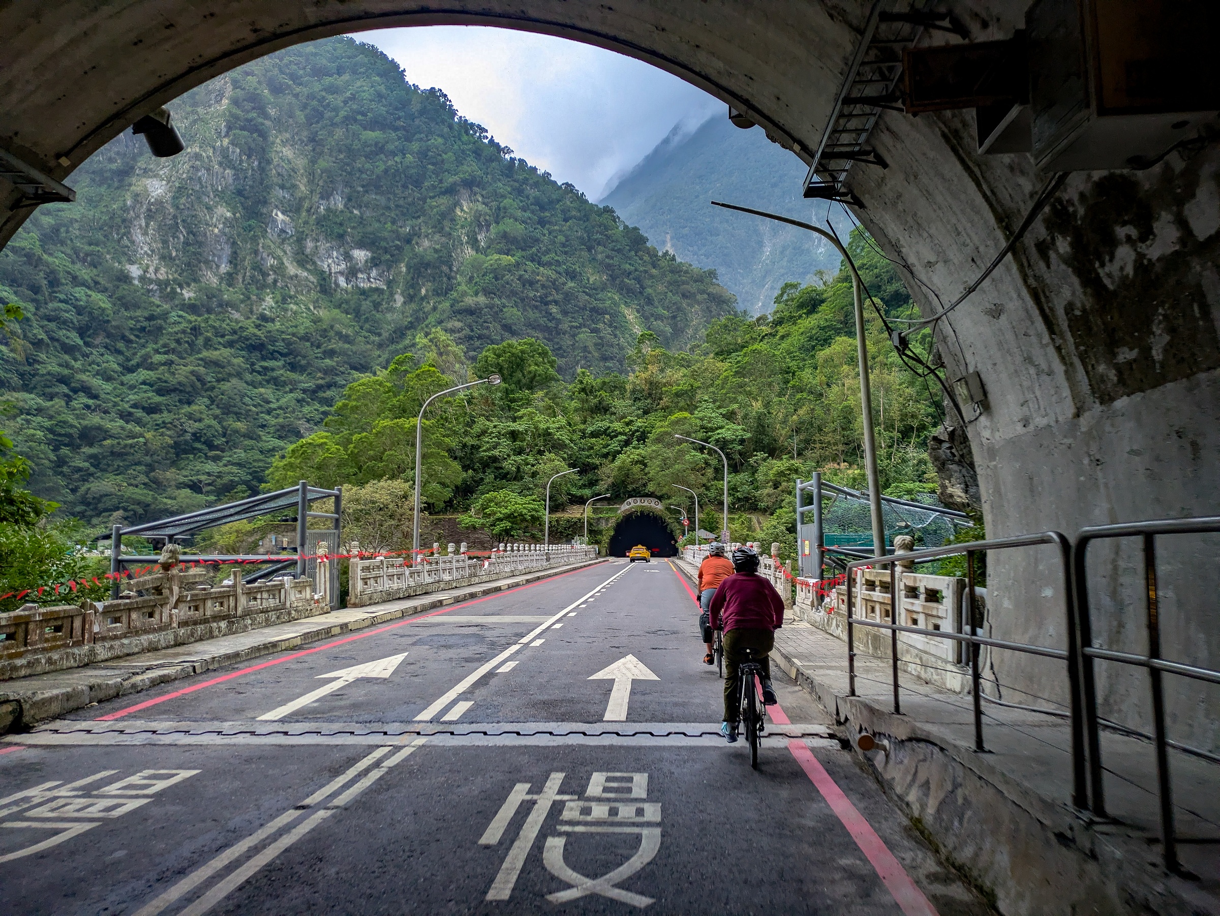 Cyclists ride along a road leading through a tunnel toward a lush, green valley.