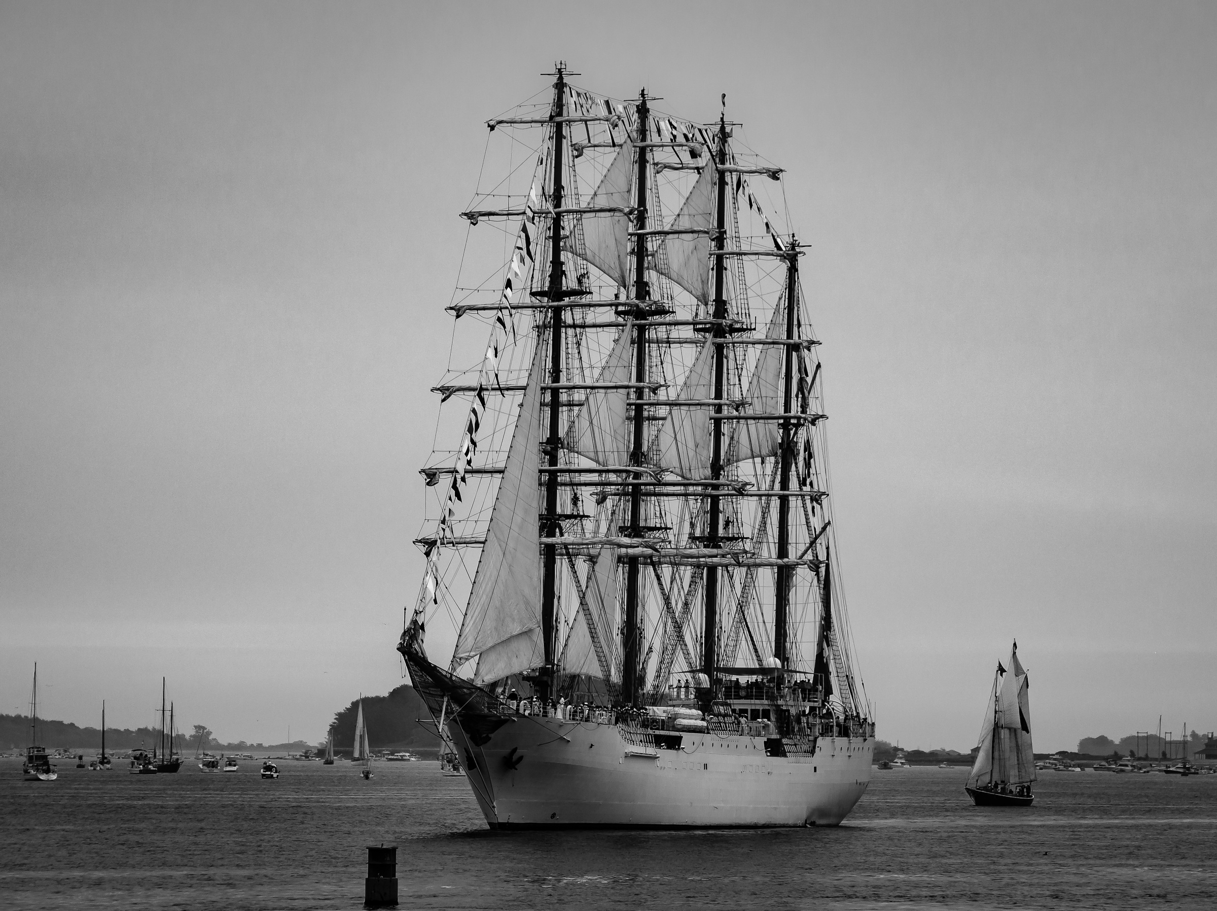A tall sailing ship floats on calm water with other boats and a distant shoreline.