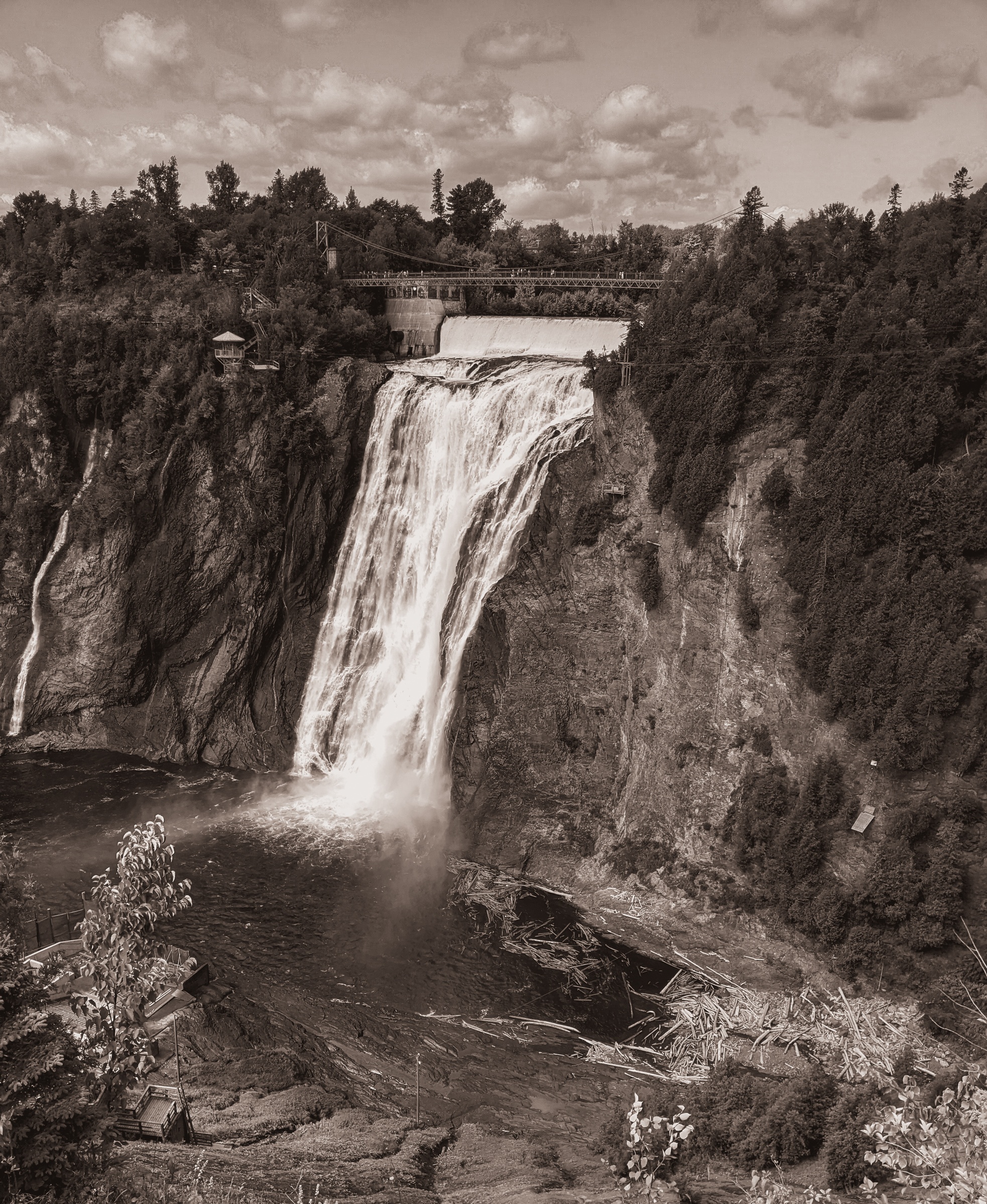 A large waterfall cascades down a rocky cliff face surrounded by trees and a historic bridge.