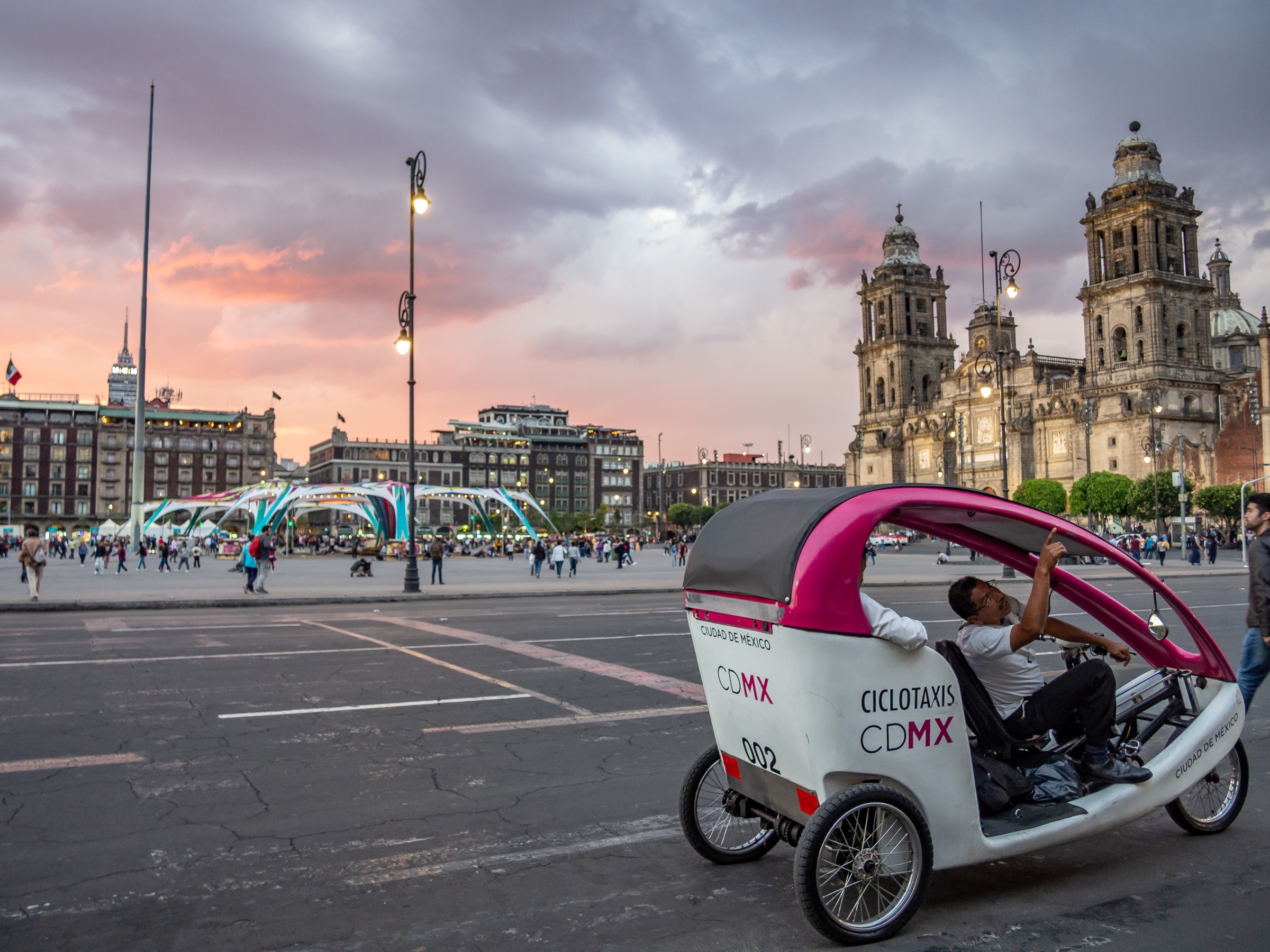 A cyclist drives a pink and white pedicab across a plaza with historic buildings and a cathedral.