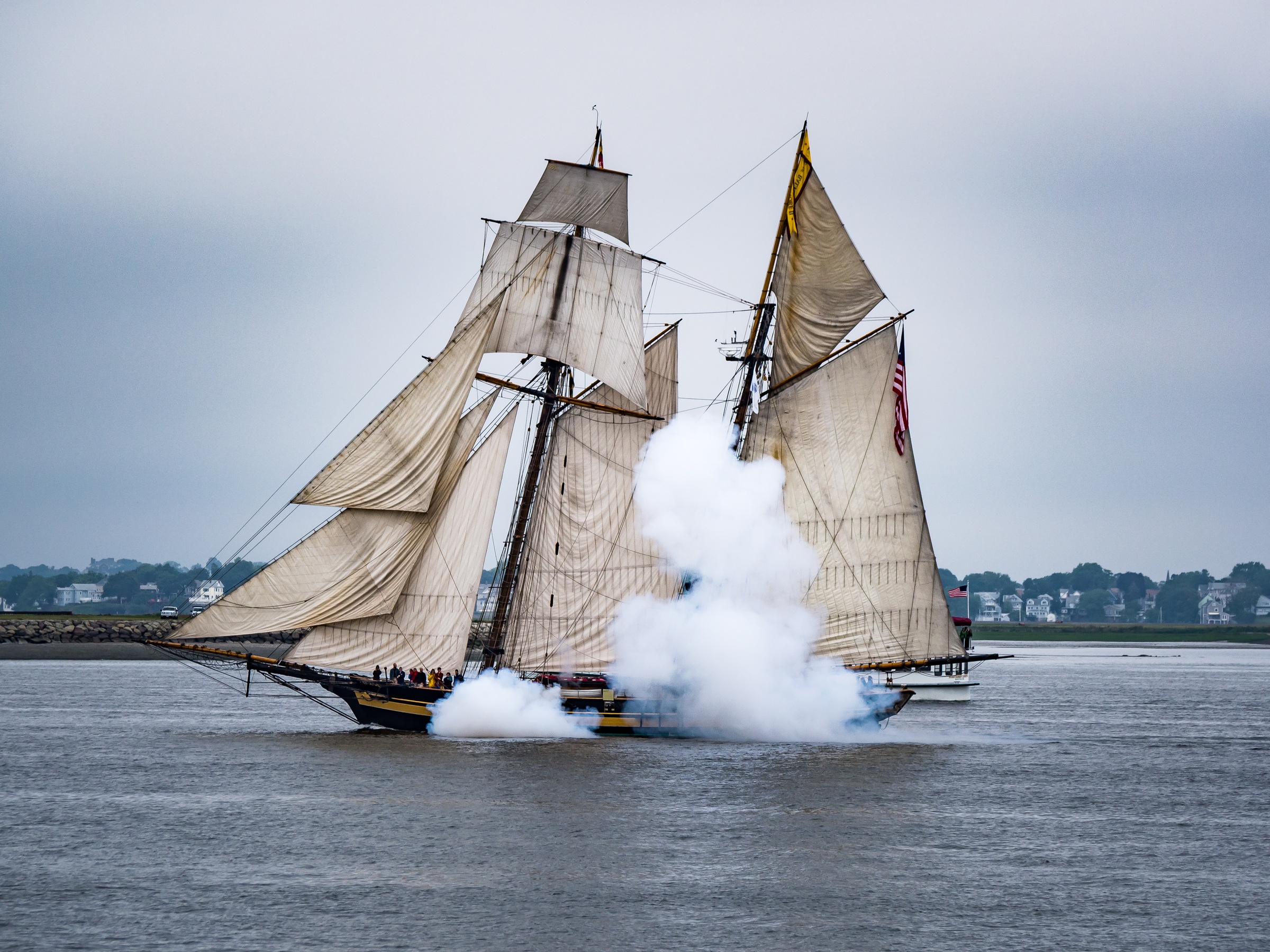 A tall ship with billowing sails fires a cannon in a river against a cloudy sky.