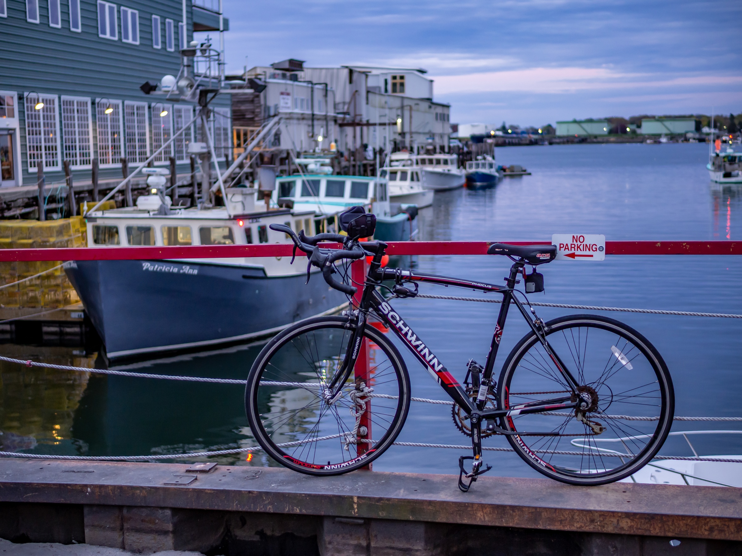 A red and black bicycle rests on a pier overlooking a harbor with fishing boats.