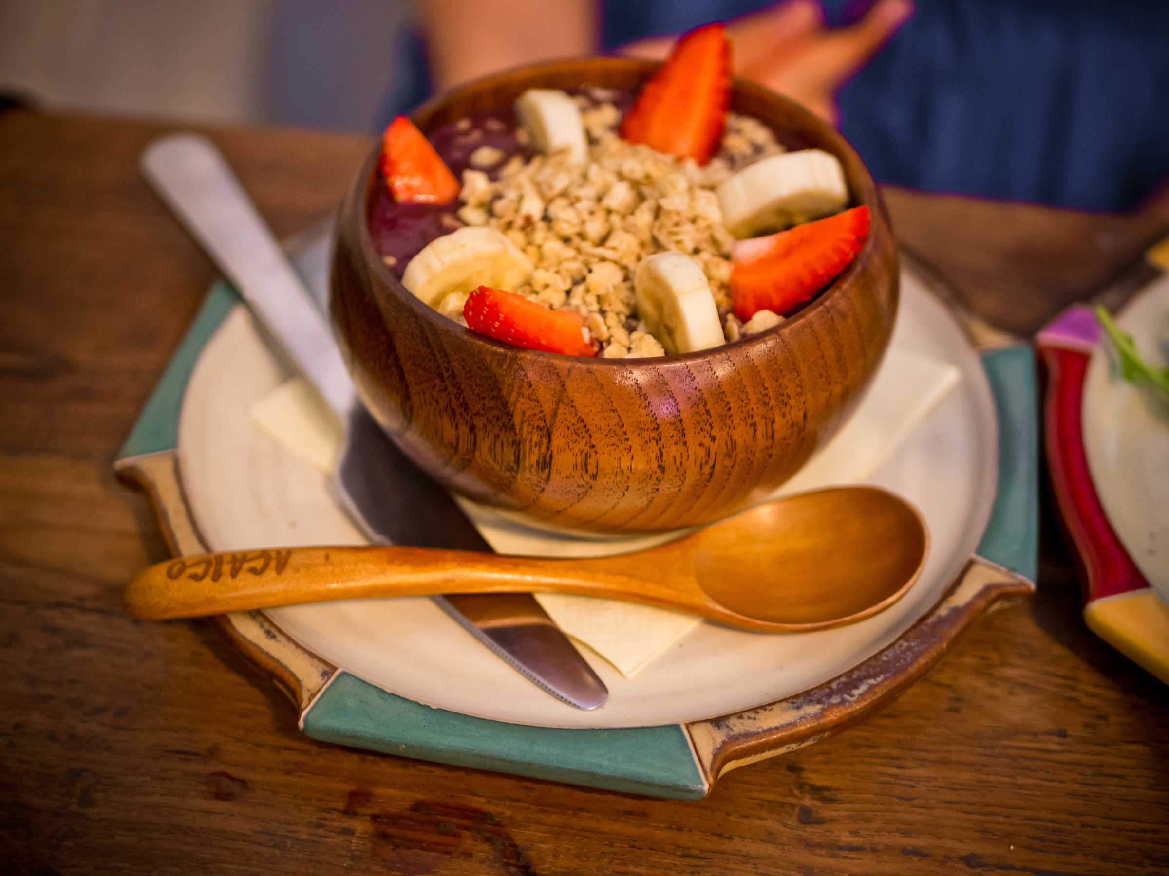 A bowl of açai with fruit and granola sits on a rustic table with a spoon and plate.