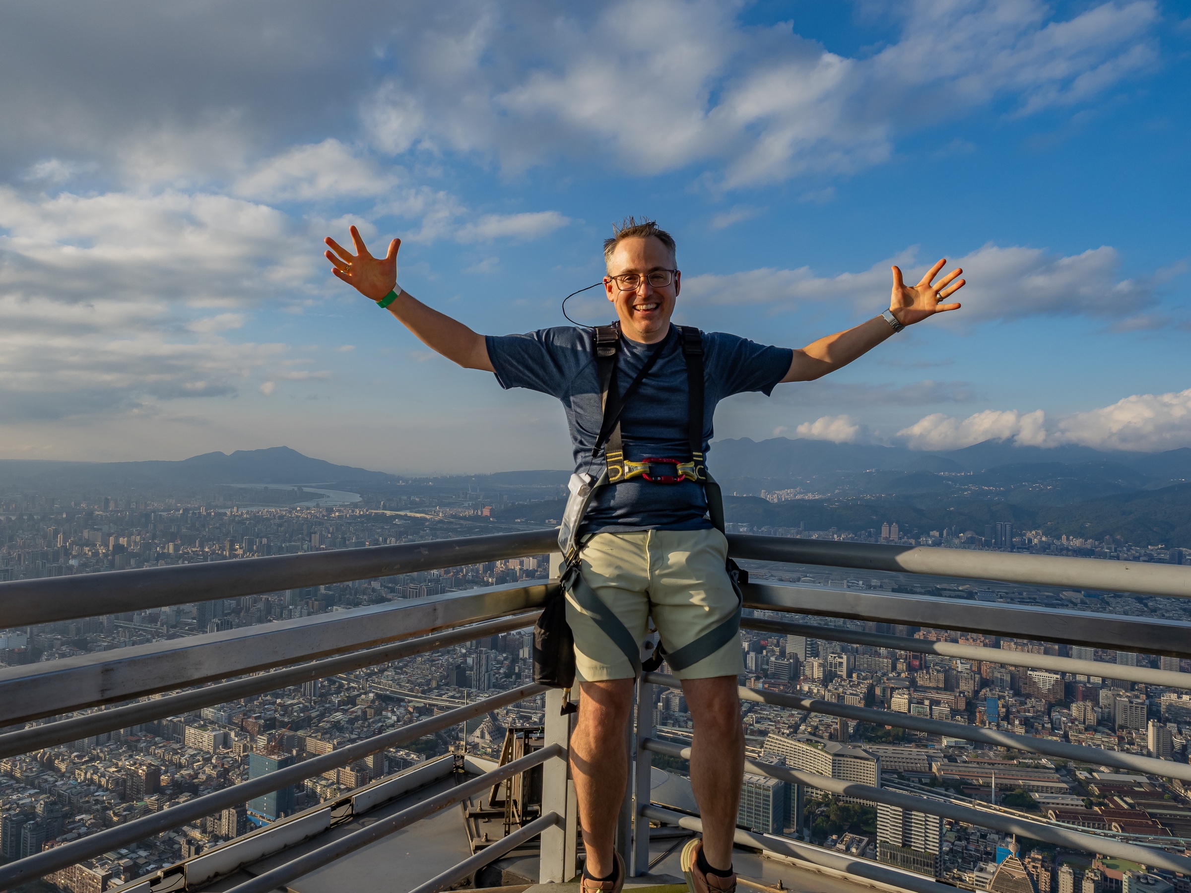 A man with arms outstretched stands on a high observation deck overlooking a sprawling cityscape.