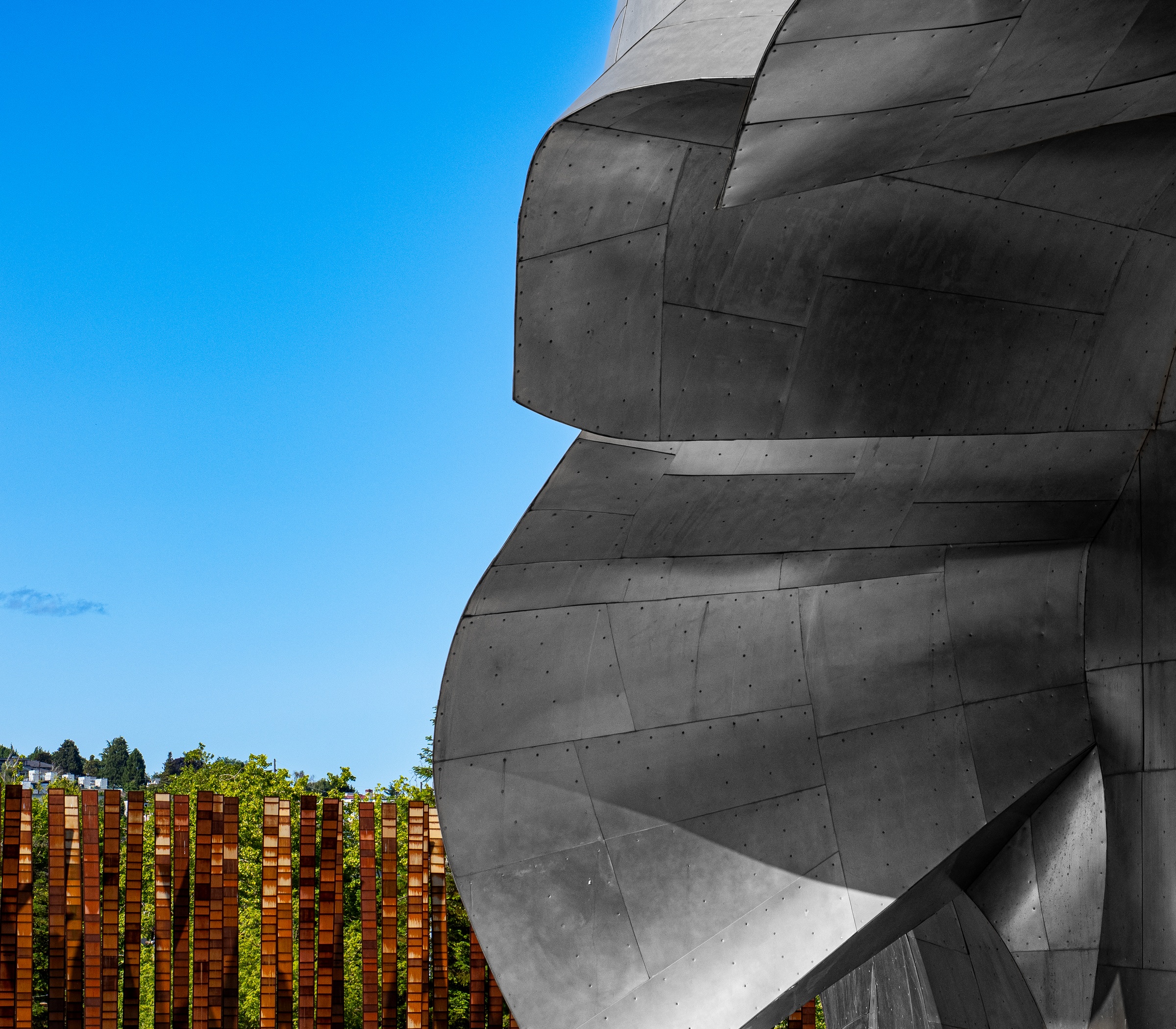 A gray, metallic sculpture sits against a bright blue sky with trees and brickwork visible below.