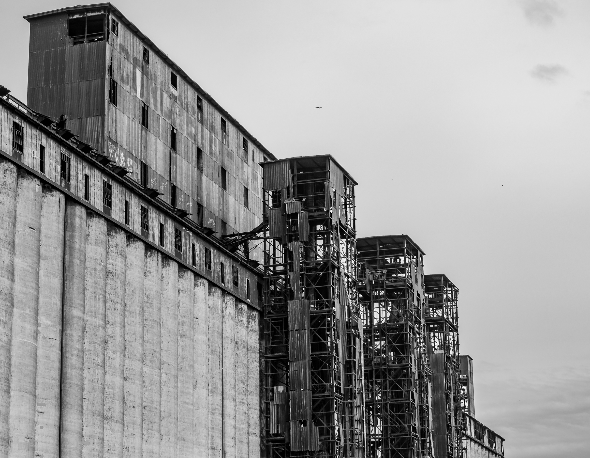 A black and white photograph shows a tall, weathered grain elevator complex against a cloudy sky.