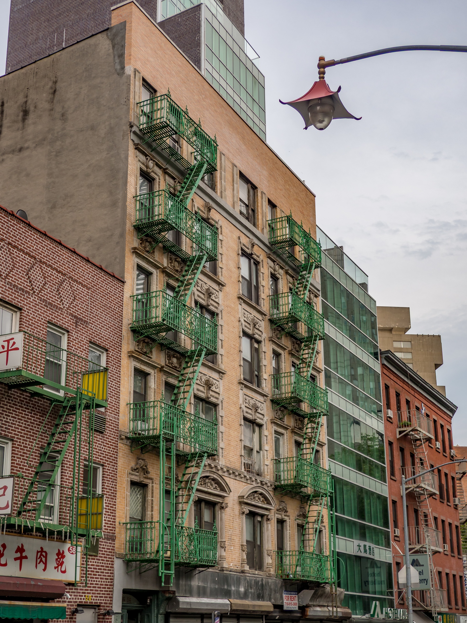 A six-story brick building with green fire escapes sits on a city street lined with other buildings.
