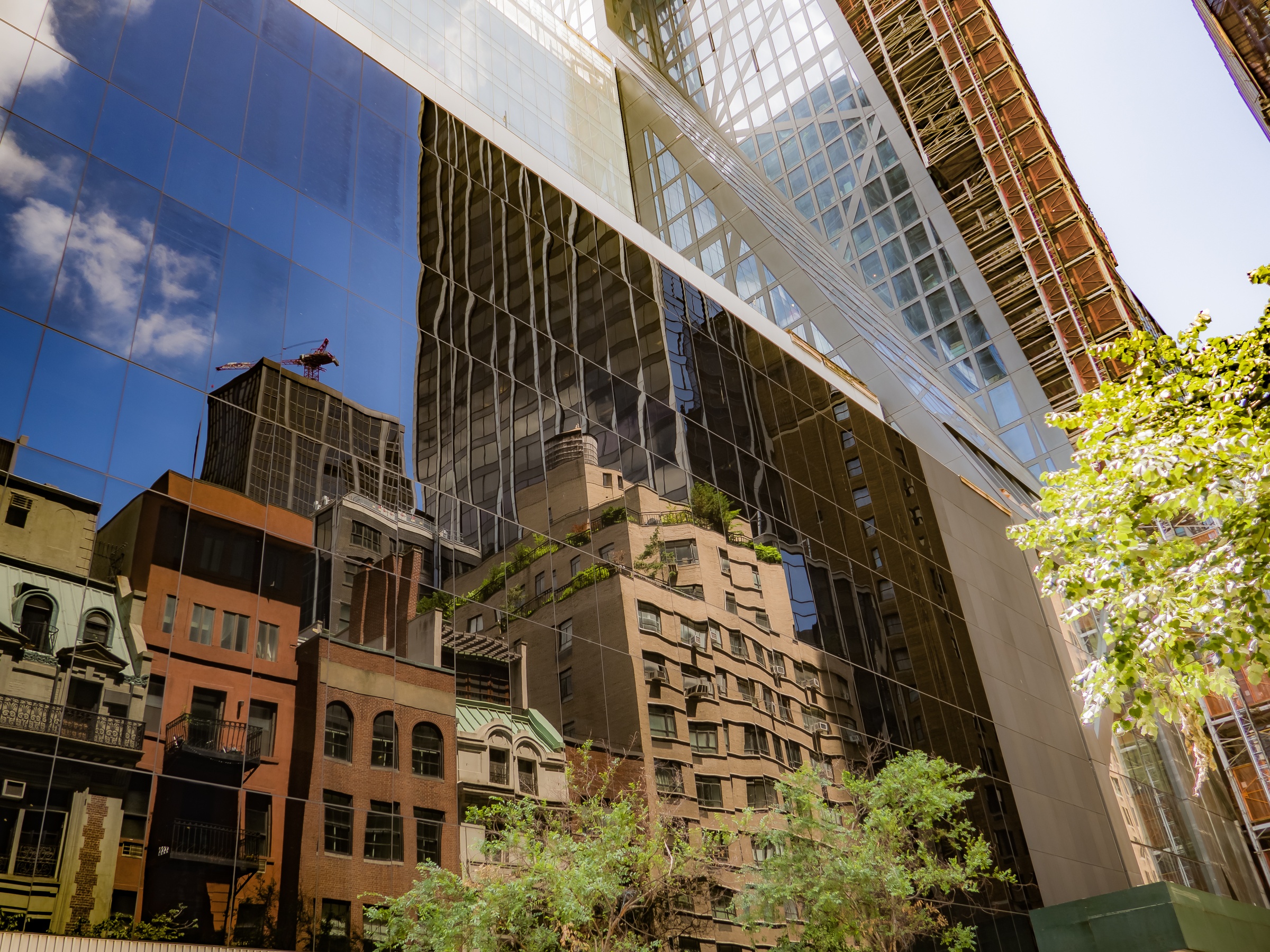 Modern glass buildings reflect older brick structures and a blue sky in a dense urban setting.