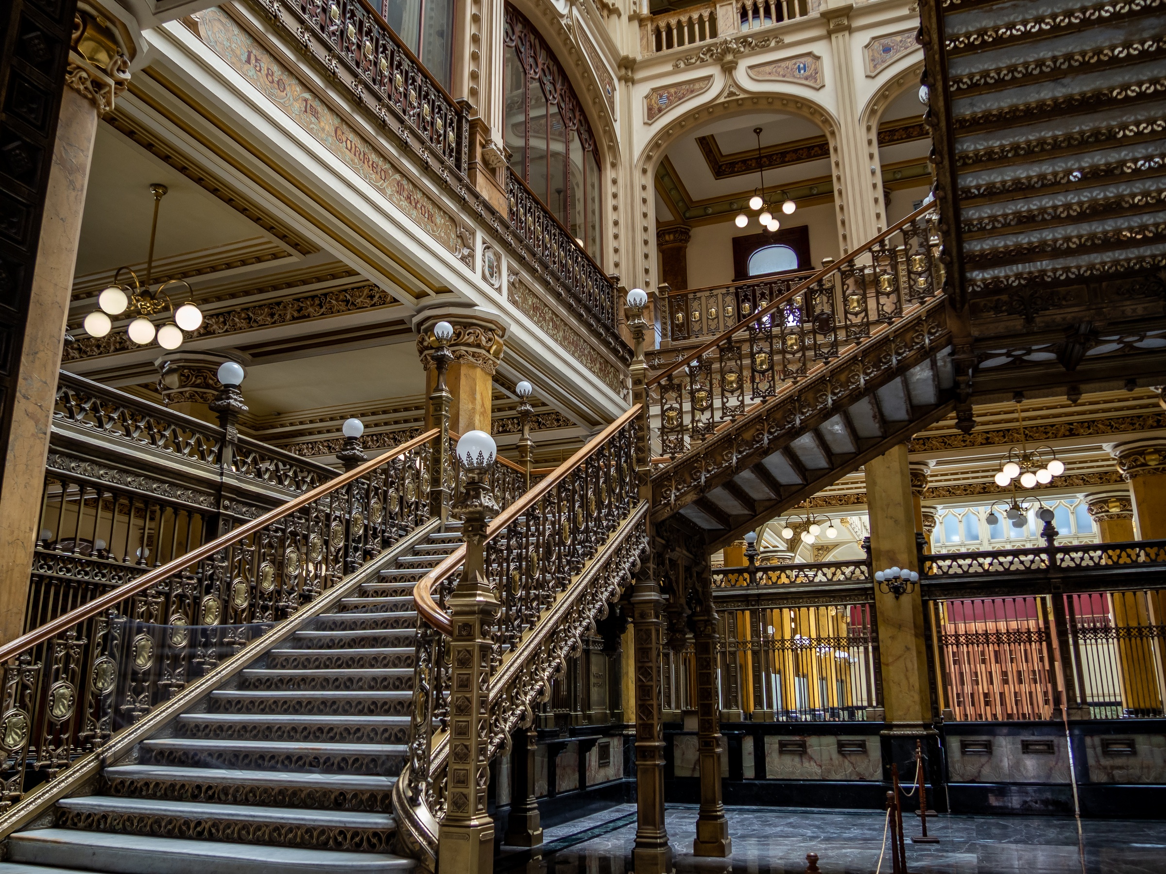 Ornate, sweeping staircases with decorative ironwork rise within a grand, historic building.