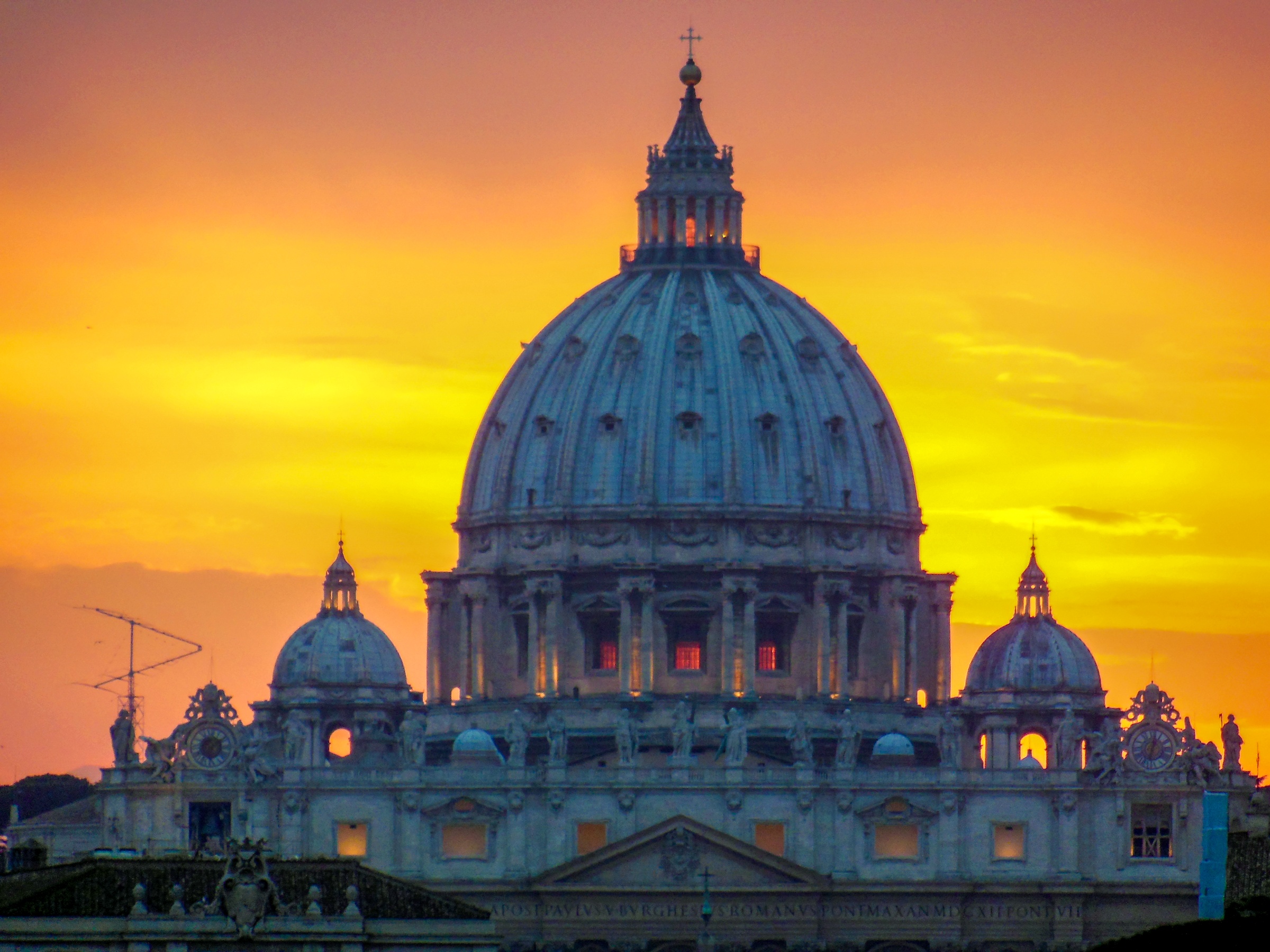 St. Peter's Basilica domes are silhouetted against a vibrant orange and yellow sunset sky.