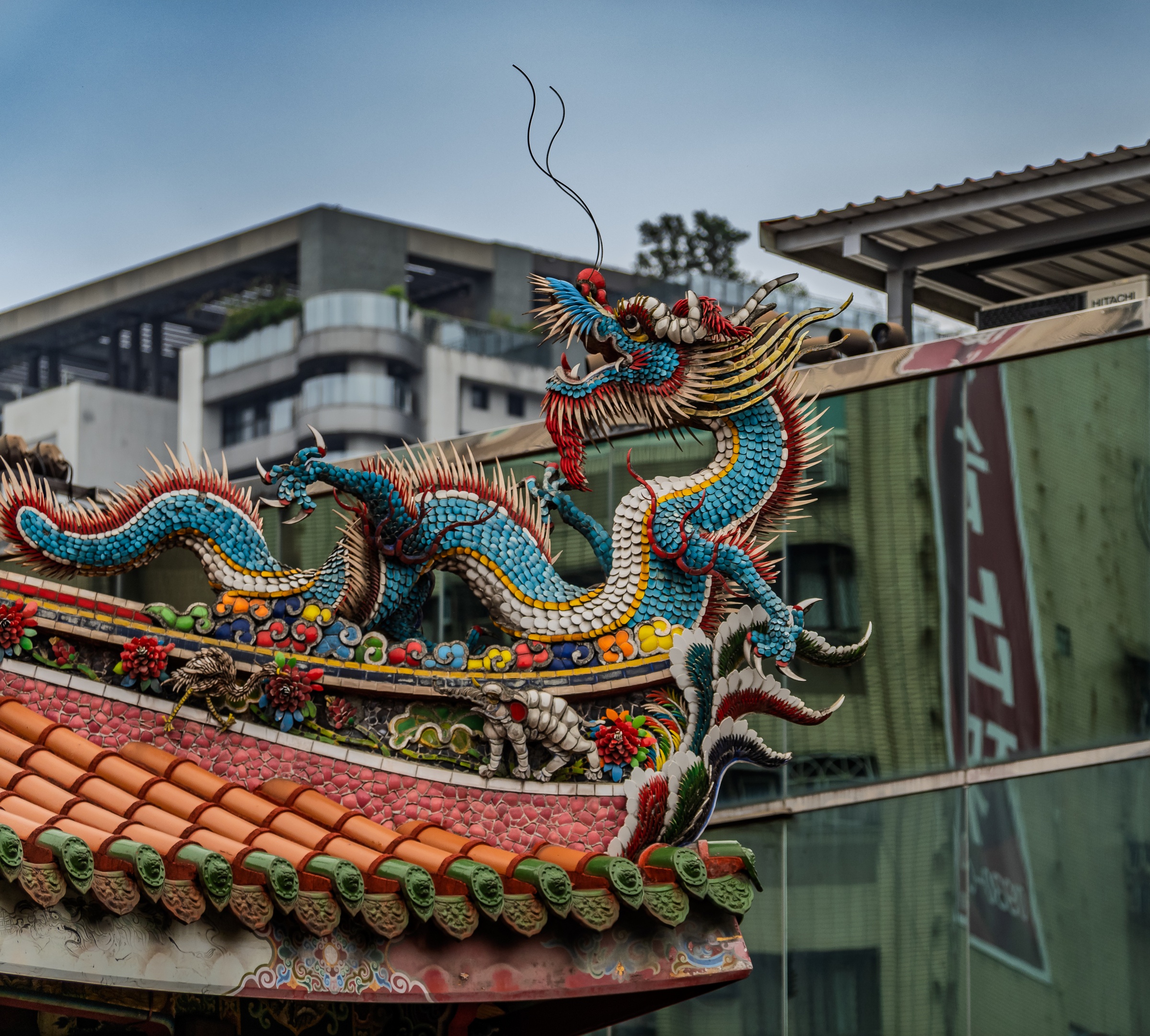 A vibrant, detailed ceramic dragon adorns a temple roof against a modern building backdrop.