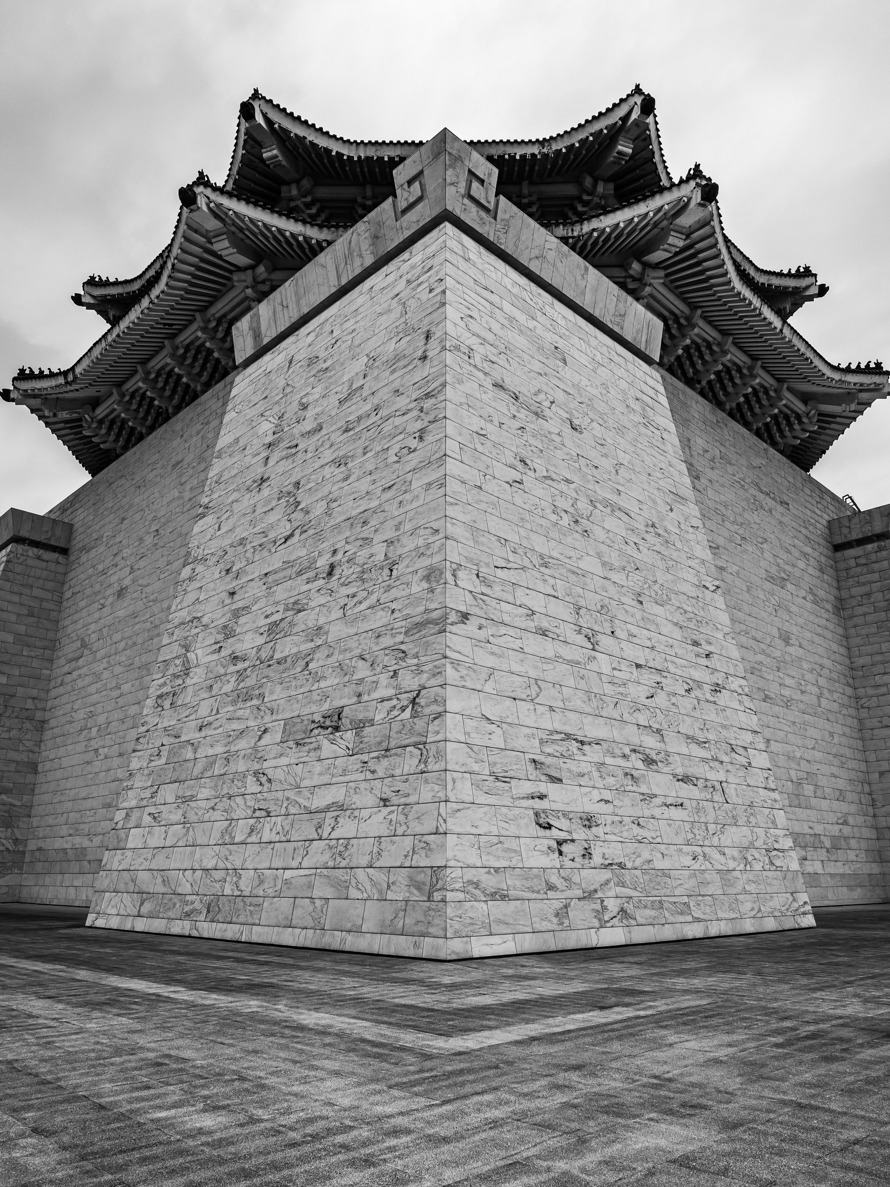A large stone building with a distinctive curved roof sits on a paved area under an overcast sky.